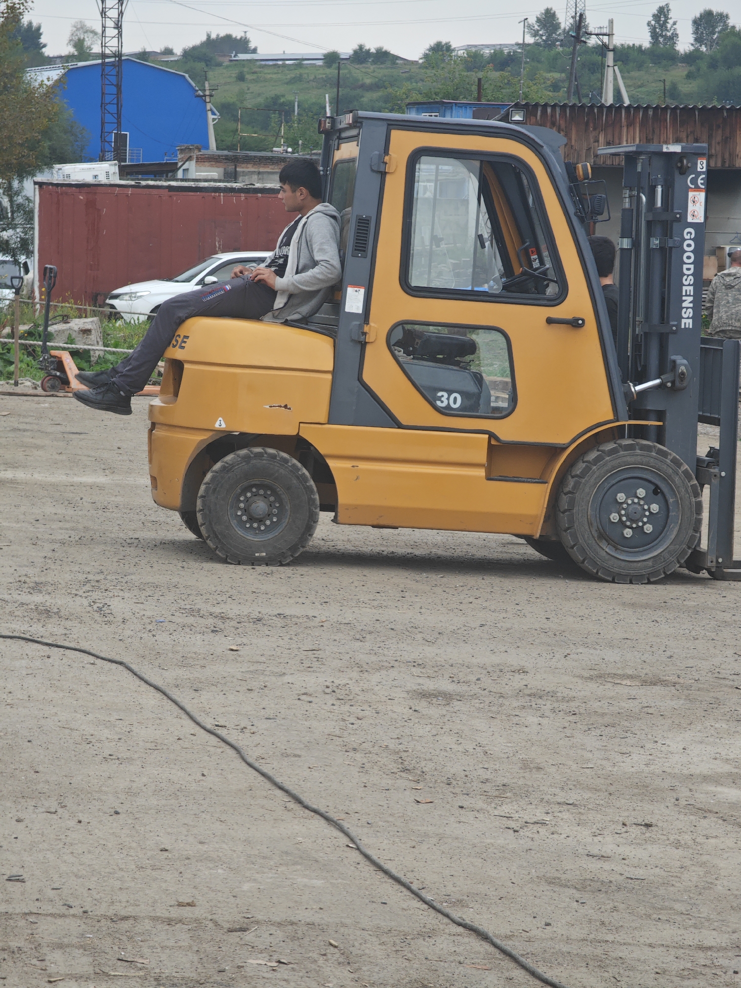 A young man in a gray hoodie and dark pants sits on the front fender of a yellow forklift. The forklift has a black cab with a window and a "30" visible on its side. The man is looking to his left, away from the camera, with his legs crossed. The forklift is on a dusty, unpaved surface, possibly a construction site or industrial yard. In the background, there are buildings, including a large blue structure, a red shipping container, and a wooden building with a corrugated metal roof. Trees and utility poles are also visible in the distance. The sky is overcast. This scene is consistent with an industrial area in Krasnoyarsk, Russia.