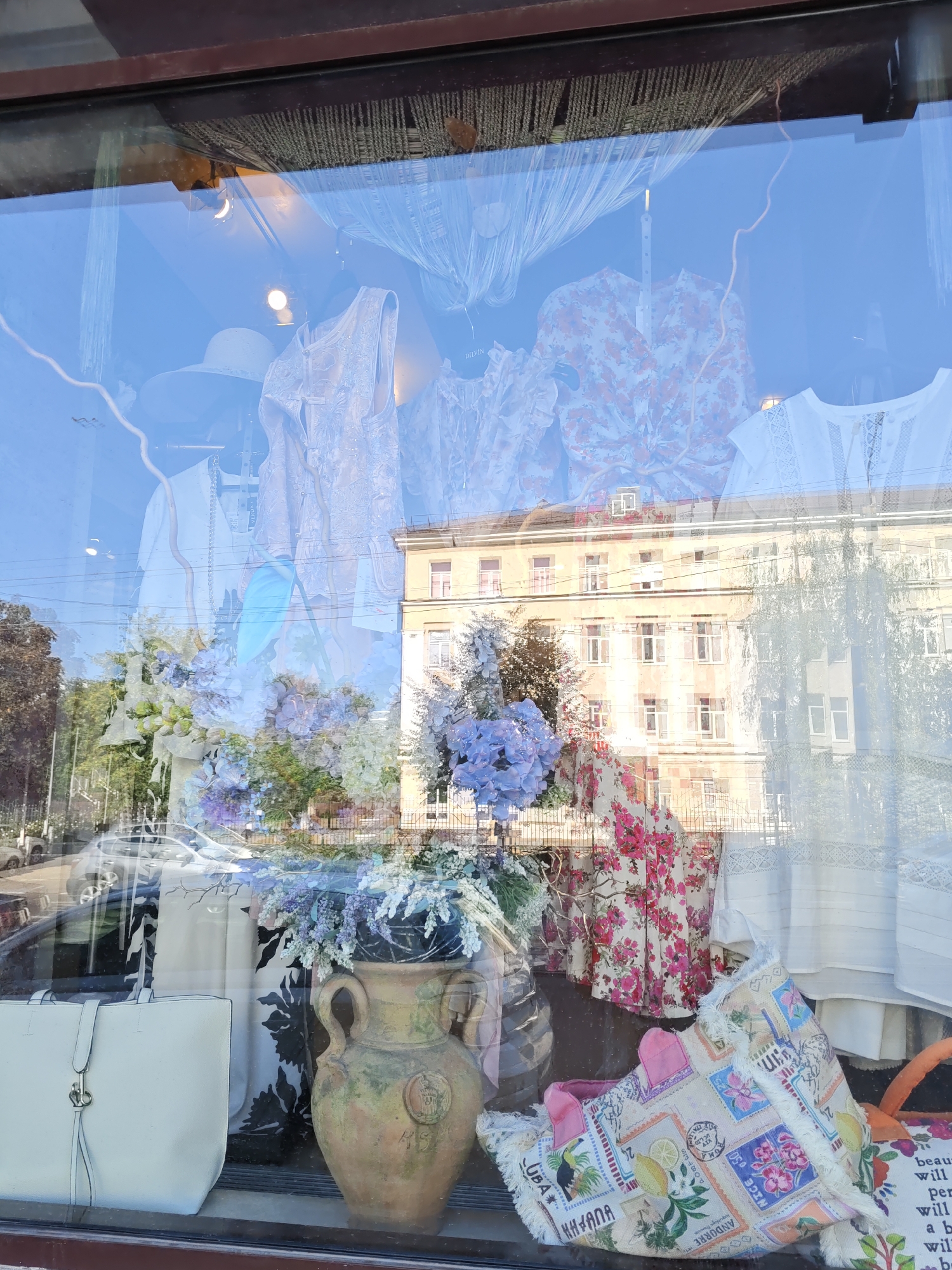 This is a storefront window in Rostov-on-Don, Russia, reflecting a street scene. The window displays various items of clothing, including white dresses and blouses, a floral print dress, and a wide-brimmed hat. There are also floral arrangements, a large ceramic vase, and two bags, one white and one with a stamp-like pattern. The reflection shows a building with multiple windows and balconies, trees, and cars. The sky is clear and blue.