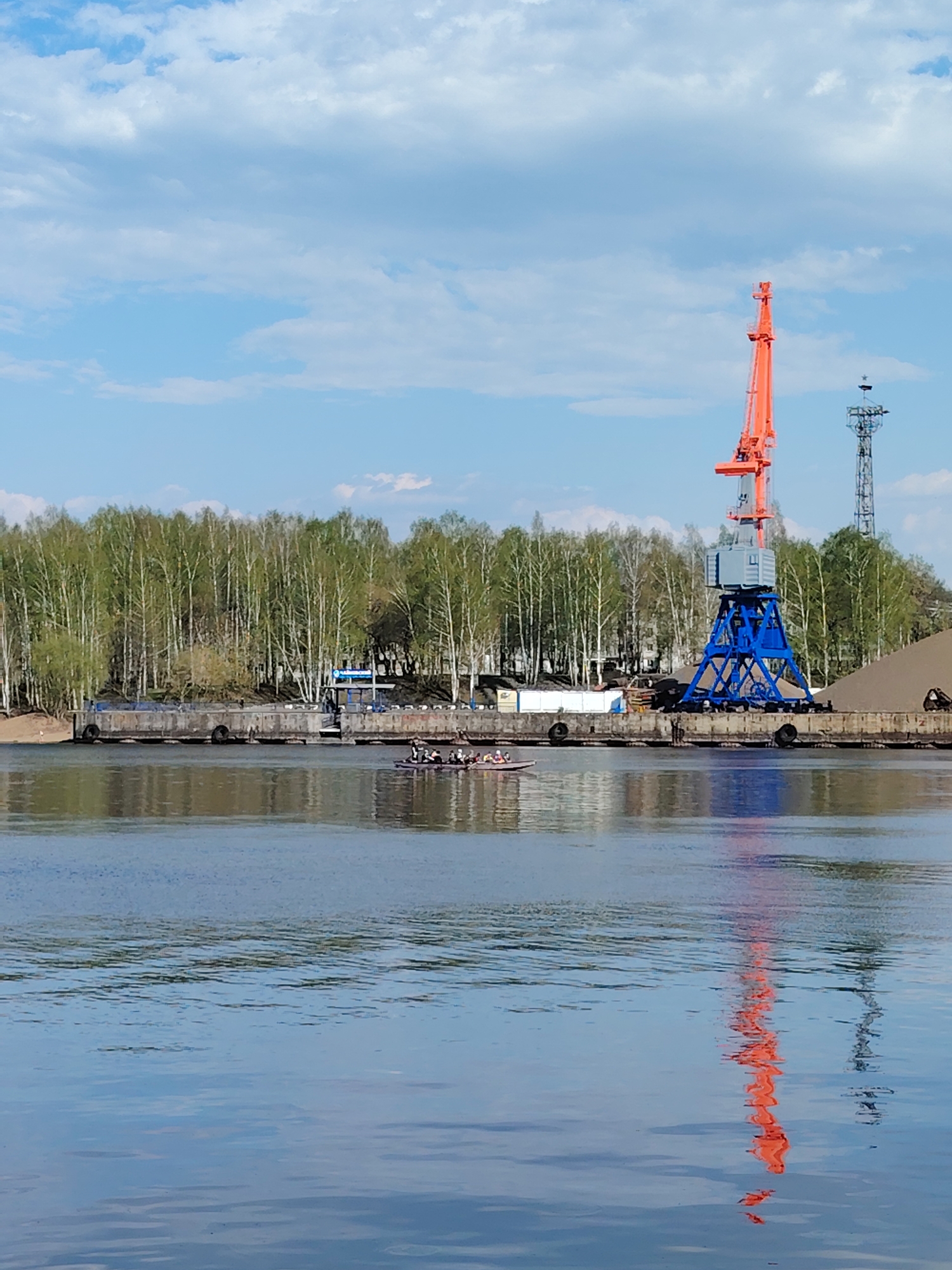 The image depicts a river scene in Chaikovsky, Russia. In the foreground is a body of water, likely the Kama River, with reflections of the crane and sky. A rowing team in a boat is visible on the river's surface. In the background, a pier with a large blue and orange crane is present. Behind the pier, a line of green trees is visible, with a telecommunications tower slightly to the right of the crane. The sky is blue with scattered white clouds.