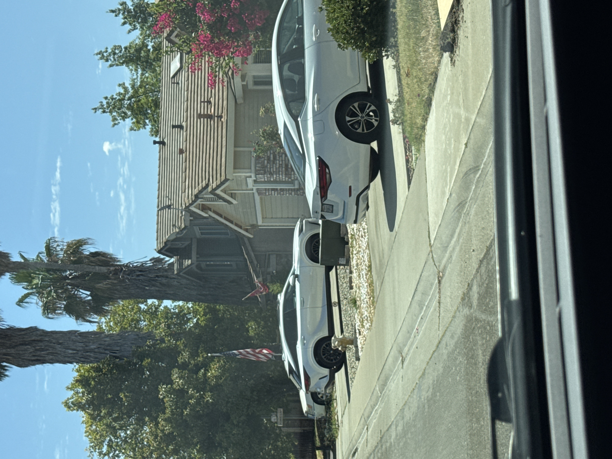 In Antelope, California, on a sunny day, a residential street is depicted with a focus on several cars parked in driveways and on the street. The foreground shows the interior of a vehicle, possibly a car, with its dashboard and windshield wipers visible. Beyond the vehicle, a white sedan and a white SUV are parked in a driveway. The SUV, a Lexus RX, is elevated on what appears to be a car lift. An American flag flies in front of the SUV. Behind the cars, a single-story house with a light-colored facade and a brown tiled roof is visible. Mature palm trees and dense green foliage surround the house, indicating a warm climate. The sky is blue with scattered white clouds. The street is paved, with concrete sidewalks and driveways. The scene suggests a typical suburban neighborhood in Antelope, California.