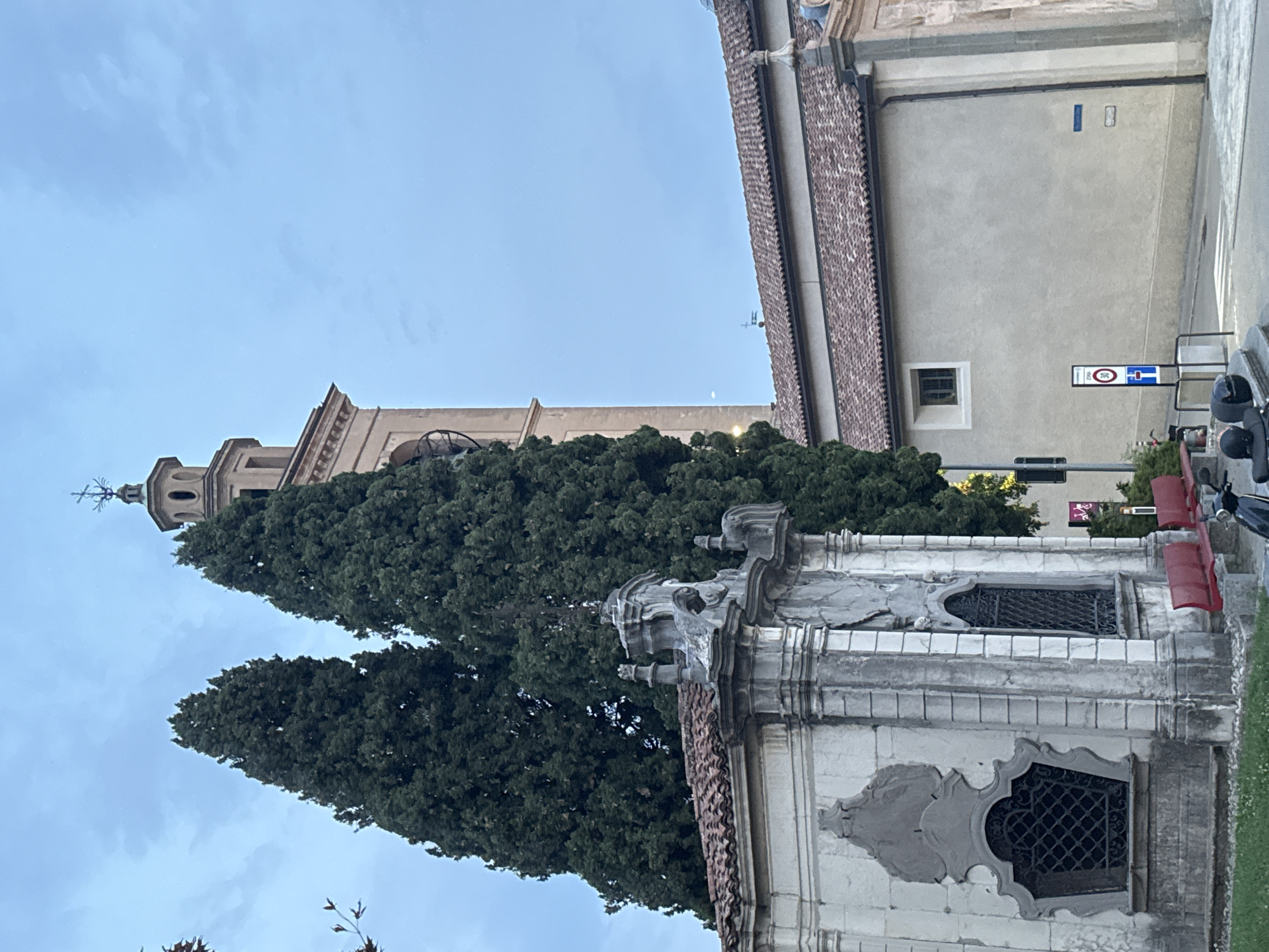This image depicts a view of the Chiesa di San Michele in Balerna, Switzerland. The foreground features the ornate stone facade of a smaller structure, possibly a chapel or mausoleum, with decorative elements and arched, barred windows. Red benches are visible in front of this structure. Behind it, two tall cypress trees stand prominently. To the right, the main church building with its tiled roof and a square window is visible. A tall, slender bell tower with a clock face rises in the background, topped with a cross. The sky is a clear, pale blue with faint cloud formations. A traffic sign indicating a speed limit of 30 is positioned near the road, and a scooter is parked in the lower right corner. The scene appears to be captured during the late afternoon or early evening.