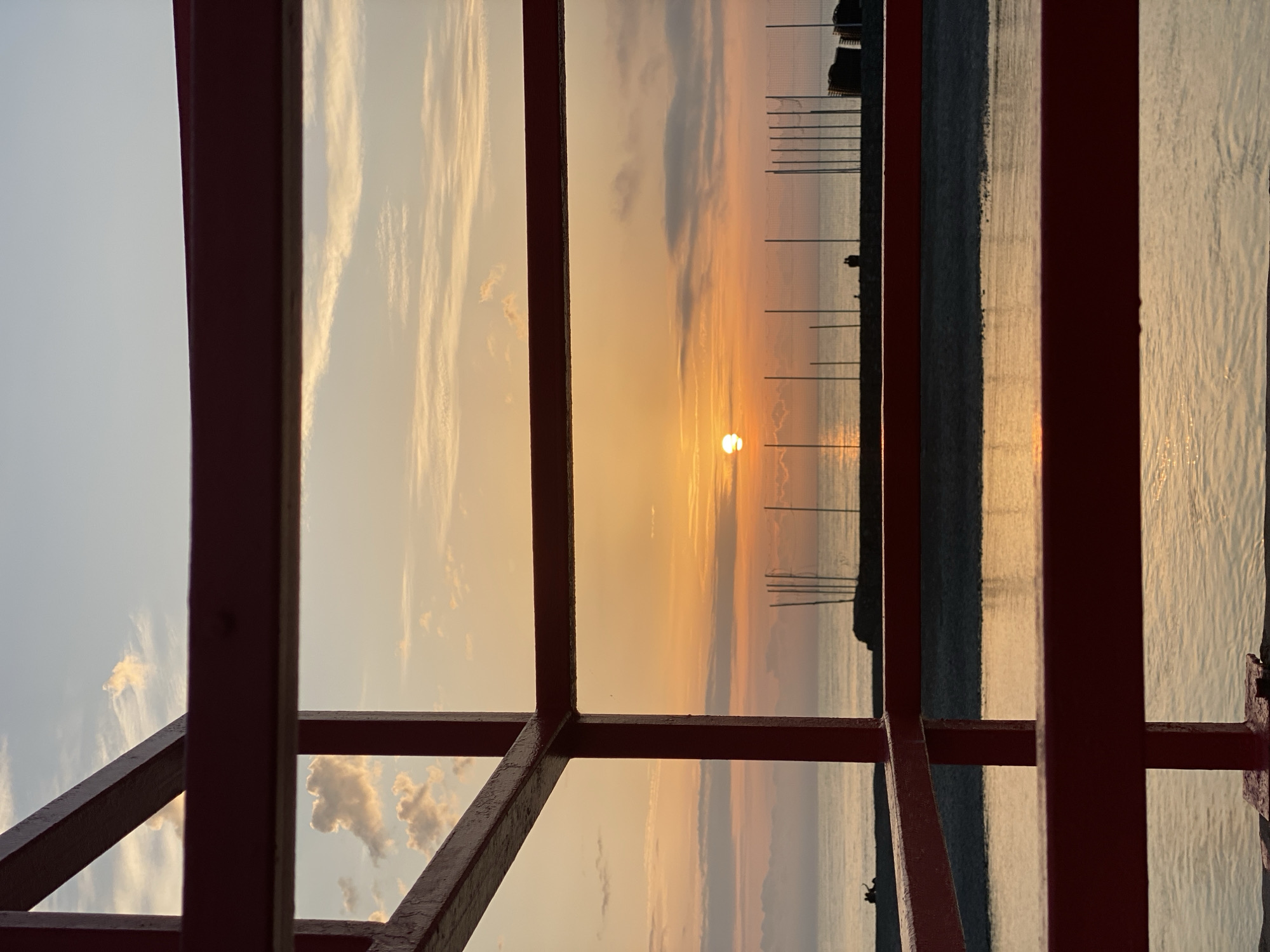 The image presents a sunset scene viewed through a red metal structure, likely a framework or scaffolding. The sunset is visible through the openings in the structure, with the sun appearing as a bright orb in the center of the frame. The sky transitions from light blue on the left to orange and yellow hues near the sun, indicating the sunset. Thin clouds are scattered across the sky.
In the distance, a body of water is visible, reflecting the colors of the sunset. Vertical poles or posts are erected along the shoreline, and structures are on the far shore.
Based on the characteristics and environment in the image, it's challenging to definitively confirm the location in Sochi, Russia, without further information.