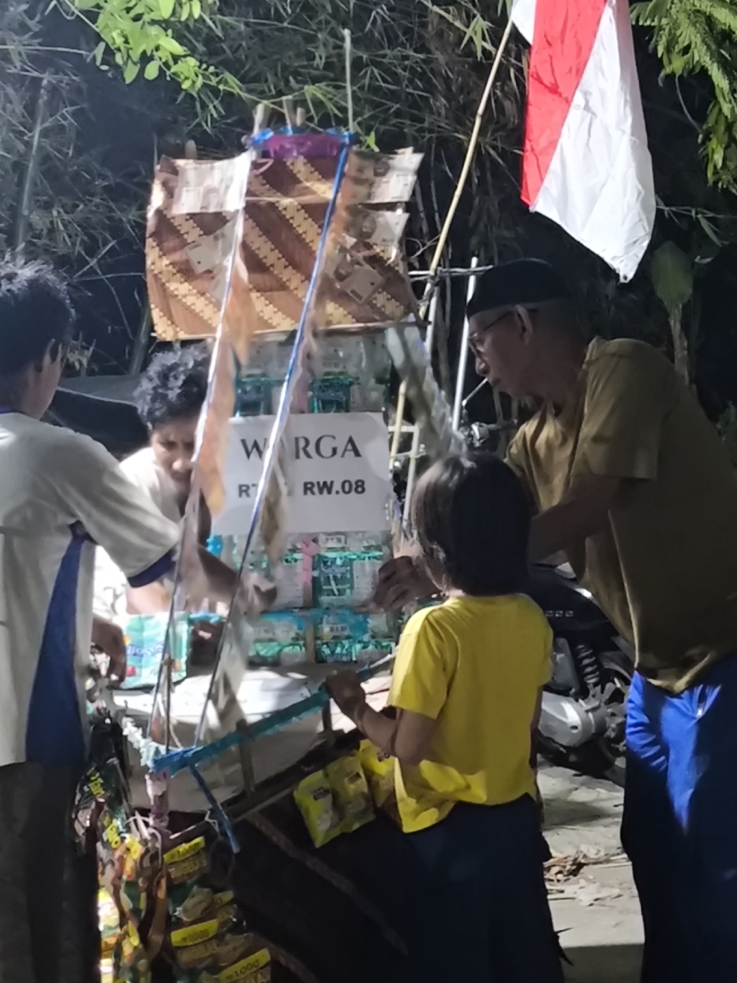 The image depicts a street scene in Cilegon, Indonesia, at night. In the foreground, a man wearing glasses and a black cap is standing next to a young child in a yellow shirt. The man appears to be interacting with the child or an object in front of them. Behind them, a red and white Indonesian flag is flying from a pole. To the left, two other individuals are visible, one in a white and blue shirt and another partially obscured. They are gathered around a stall displaying various items, possibly snacks or drinks, with some signage that reads "WARGA RT. RW.08". The background is dark with some foliage and bamboo visible, suggesting an outdoor environment. A motorcycle is parked behind the man and child. The overall scene suggests a local community gathering or market stall operation.