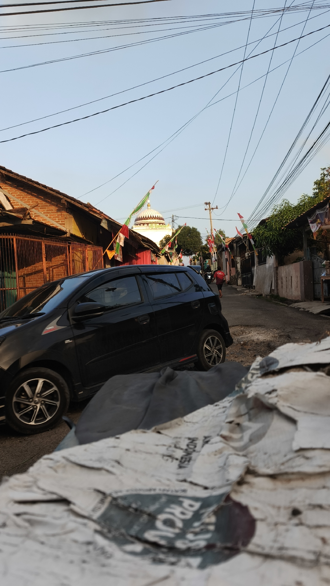 A black hatchback is parked on a narrow street in Bandar Lampung, Indonesia, in front of a building with a red facade and a metal fence. The street is lined with houses and overhead utility wires crisscross the sky. Small triangular flags in red and white are strung across the street, suggesting a celebration or national holiday. In the background, a mosque with a white dome is visible. A person in a red shirt is walking away from the camera down the street. The foreground is occupied by a pile of torn paper, some of which appears to be a map or poster with Indonesian text. The lighting indicates it is late afternoon or early evening.