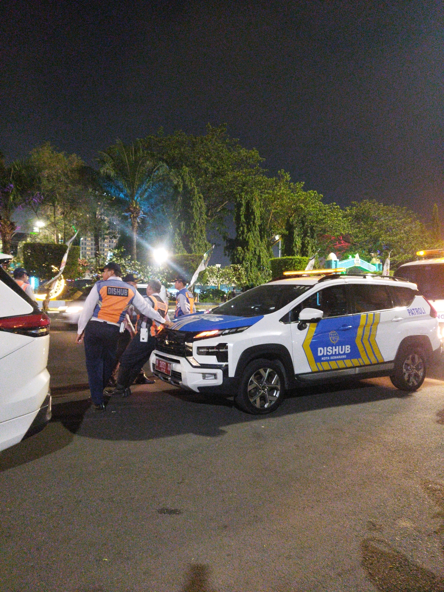 The image depicts an outdoor scene at night, likely in a city environment in Semarang, Indonesia, with several individuals and vehicles present. A white SUV with "DISHUB KOTA SEMARANG" and "PATROLI" markings on its side and "DISHUB" on its back is the prominent subject. The vehicle has a light bar on its roof and appears to be engaged in some activity, with a few people gathered around its front.
There are at least four individuals visible, all wearing similar orange vests with "DISHUB" printed on the back, over dark clothing. Two individuals are interacting with the front of the SUV, and another appears to be walking away from it. The ground is paved asphalt. In the background, there are trees, streetlights, and what appear to be buildings or structures, suggesting an urban setting. The overall lighting suggests it is nighttime, with artificial lights illuminating the scene. There is also a white vehicle partially visible on the left side of the frame. The overall impression is one of official activity or enforcement related to traffic or public transportation, given the "DISHUB" (Department of Transportation) markings.