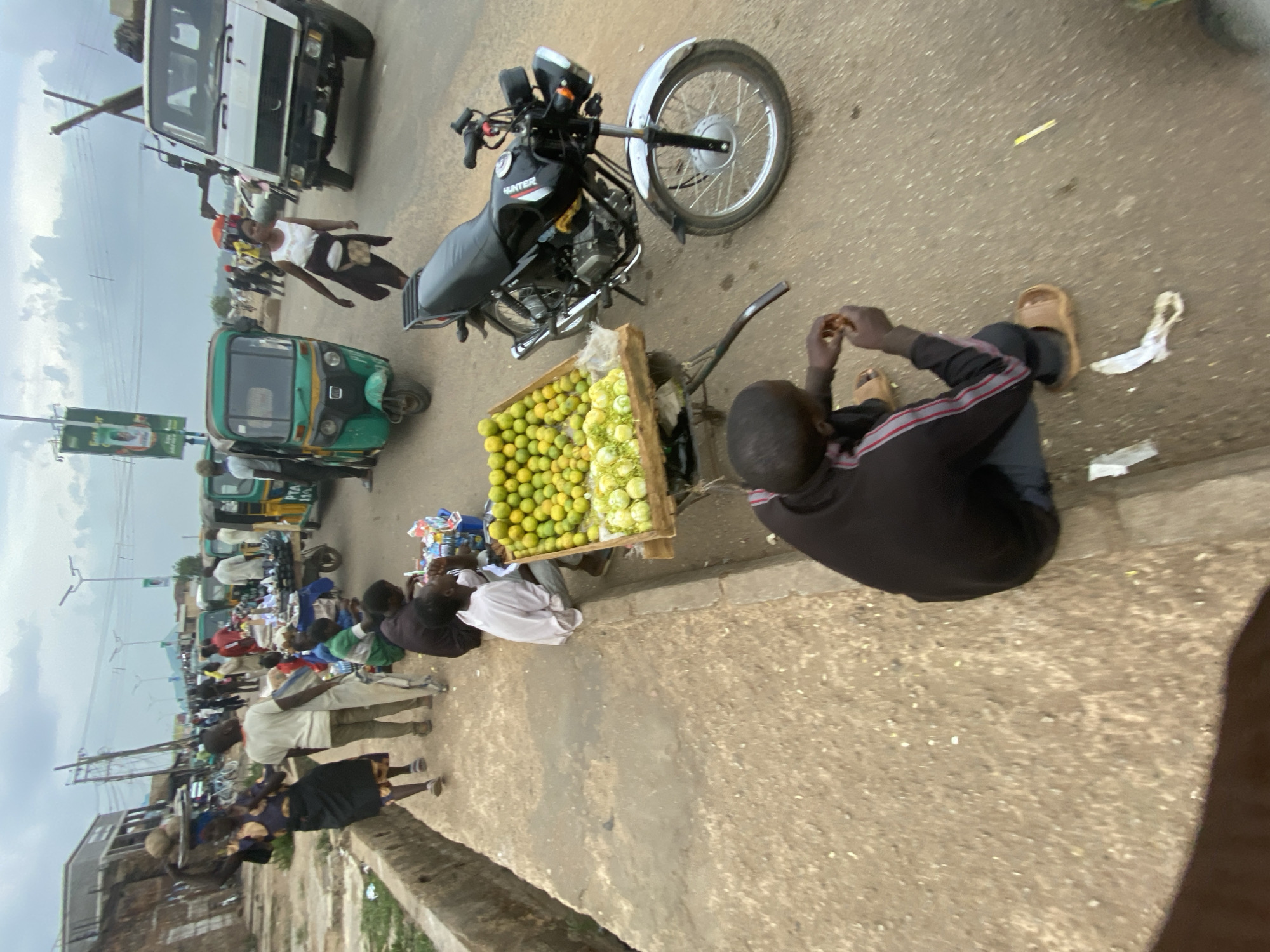 The image depicts a street scene in Bukuru, Nigeria. A man sits on the edge of a sidewalk, wearing a dark long-sleeved shirt, jeans, and sandals. He appears to be eating or handling something with his hands. Next to him is a cart filled with green and yellow fruit, likely oranges or tangerines. The cart is attached to a black motorcycle parked on the side of the road.
Along the street, there are several other people, including some standing around other vendor carts or stalls. A green auto rickshaw (tuk-tuk) is parked nearby. A white truck is visible further up the street. The sky is overcast. The scene suggests a busy street market or roadside vending area in Bukuru.