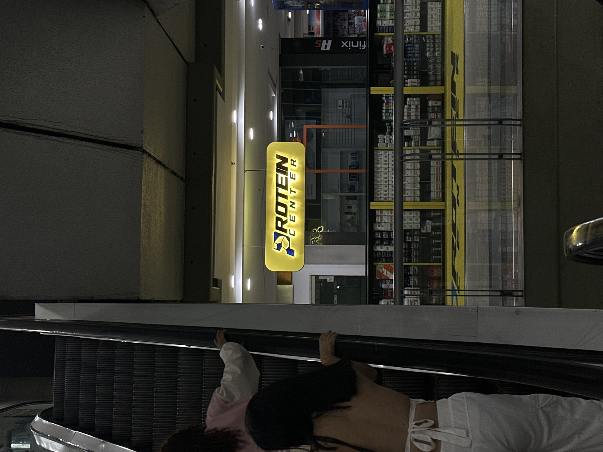 The image shows two individuals lying prone on an escalator in what appears to be a shopping mall or commercial space. One person is wearing light-colored pants and a dark top, exposing their midriff. The other person is partially visible, wearing light pink and white clothing. They appear to be positioned head-to-toe on the moving escalator.
Behind the escalator, there is a "Protein Center" sign illuminated in yellow. A shop with shelves stocked with goods is visible, possibly indicating a retail setting. The architecture suggests a modern commercial building with fluorescent lighting.
The scene is most likely in the city of Caracas, Venezuela.