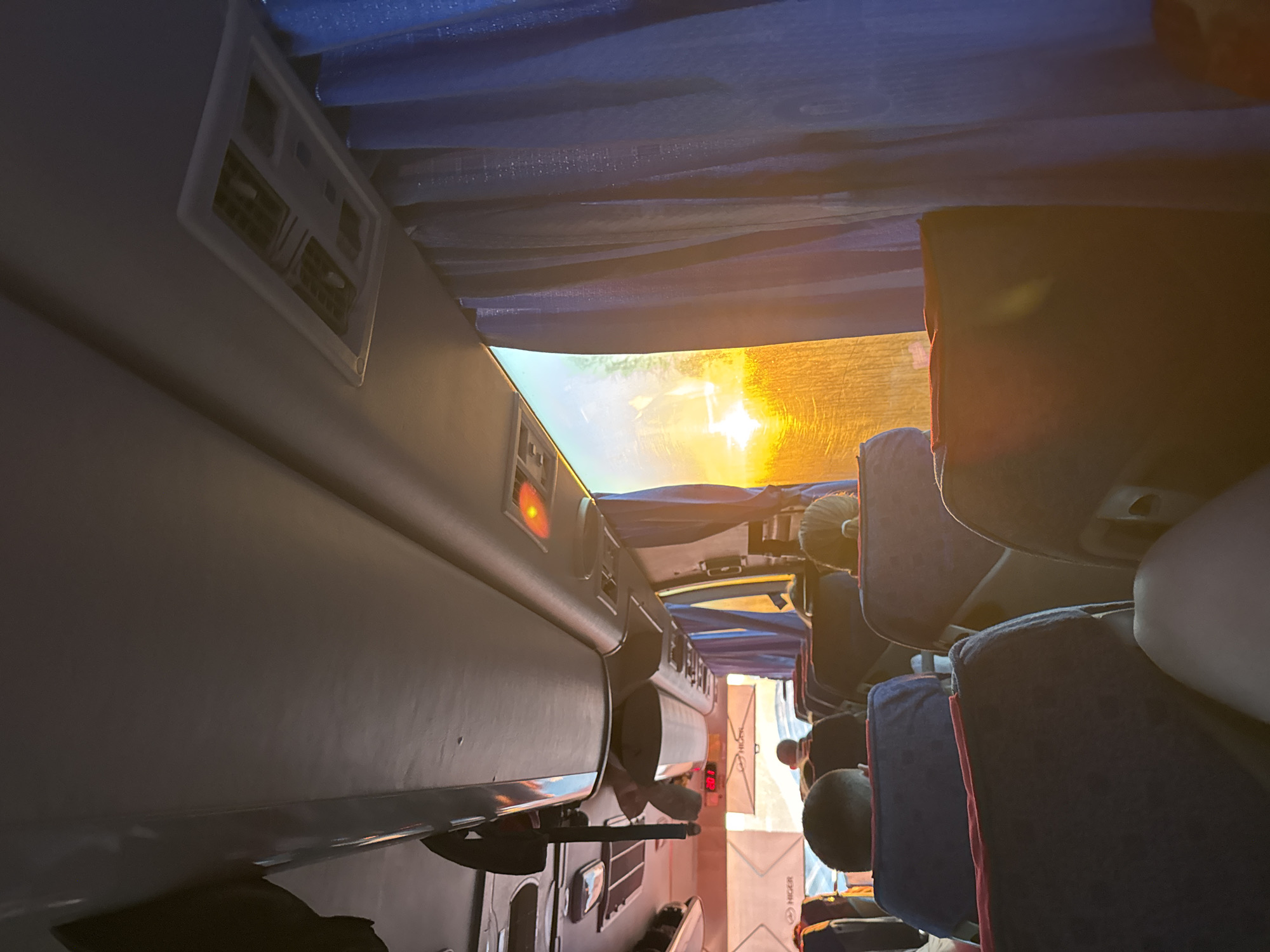 The image depicts the interior of a bus. The viewer is positioned in an aisle seat, looking toward the front of the bus, as the image has been rotated. Seats upholstered in dark blue with red trim are visible on the right side of the aisle. Passengers' heads are visible between the seats. On the left side, the wall of the bus includes vents, lights, and an emergency exit window. Bright sunlight streams through the front window, partially obscured by blue curtains. The brand name "Higer" is displayed on a placard on the front of the bus. The location, Ustinovo, Russia, is not explicitly supported by elements in the image.