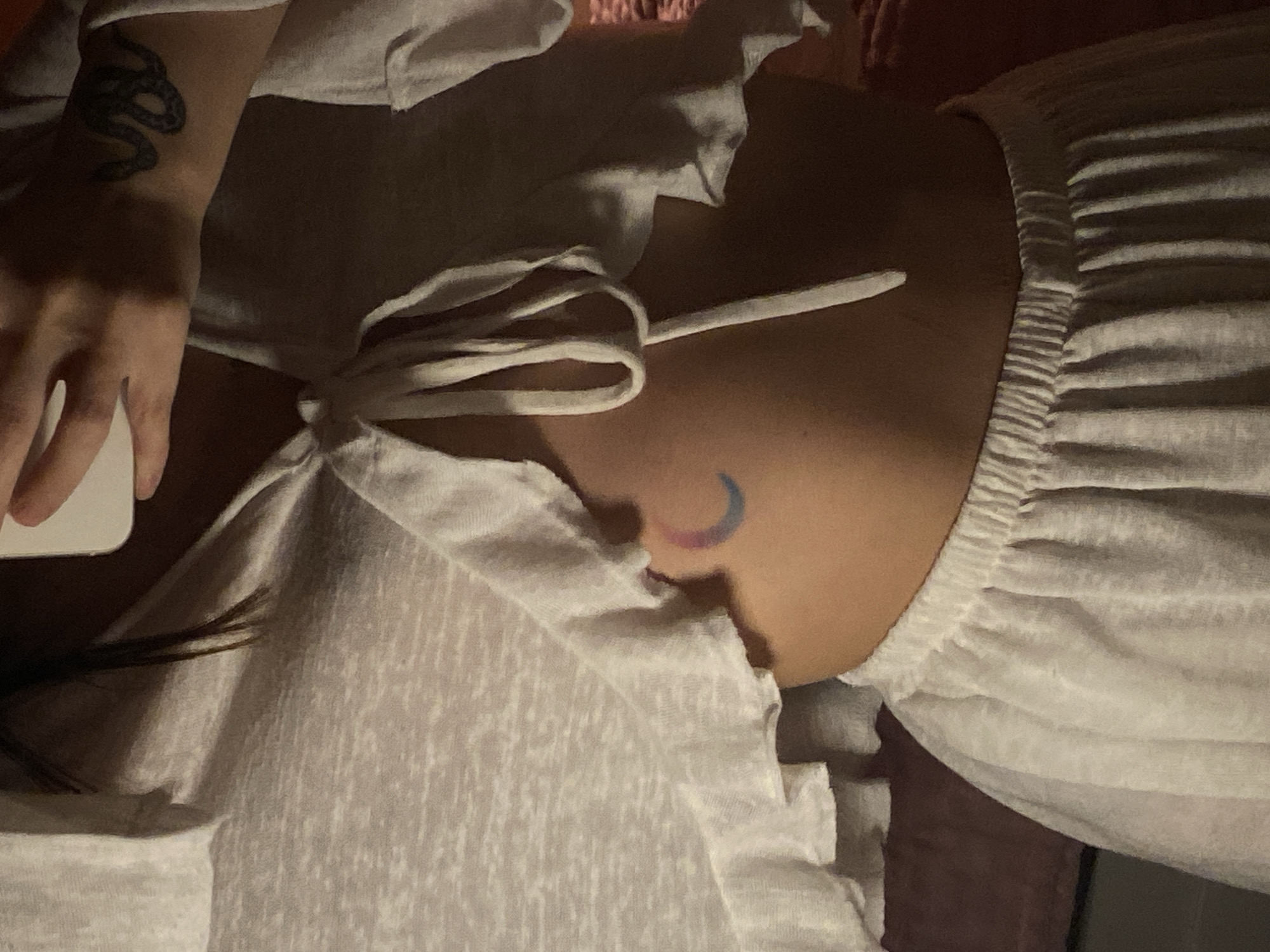 A person is seen in a low-lit indoor setting. They are wearing a white, ruffled top with drawstring detailing. On their left wrist is a snake tattoo. On their upper torso, visible under the top, is a small crescent moon tattoo with pink and blue hues. A smartphone is partially visible in their left hand. The exact location in Votkinsk, Russia is not discernible from the image.