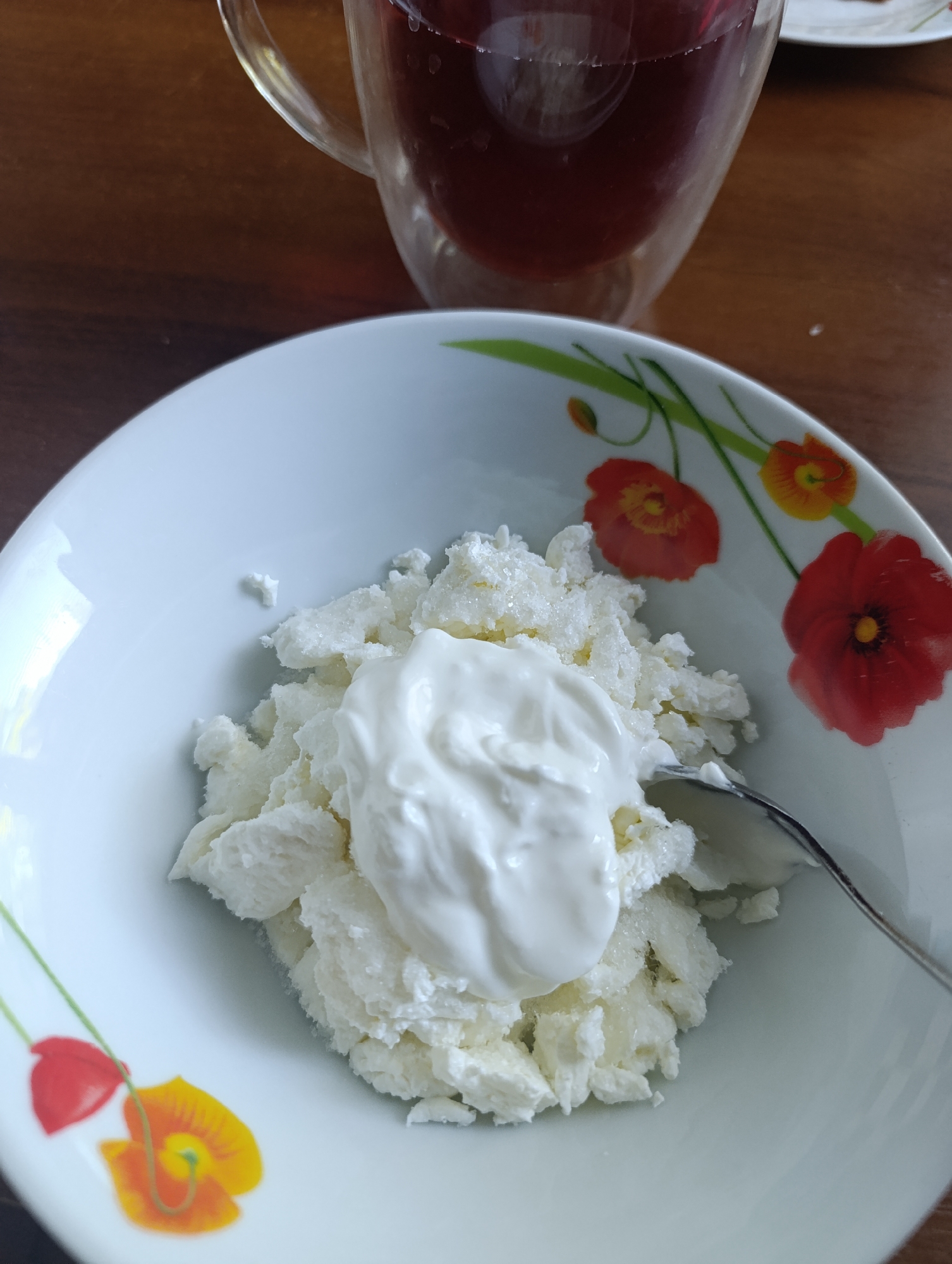 A bowl of cottage cheese topped with a dollop of sour cream sits on a wooden table. The white bowl is decorated with a pattern of red and yellow poppies. A spoon rests in the bowl, partially submerged in the cottage cheese. In the background, a glass mug filled with a dark red beverage is visible. The scene appears to be indoors, likely in a home setting, given the presence of a wooden table and a casual meal presentation. The lighting suggests it could be daytime, with natural light contributing to the scene's brightness. The location is Unknown, Unknown.