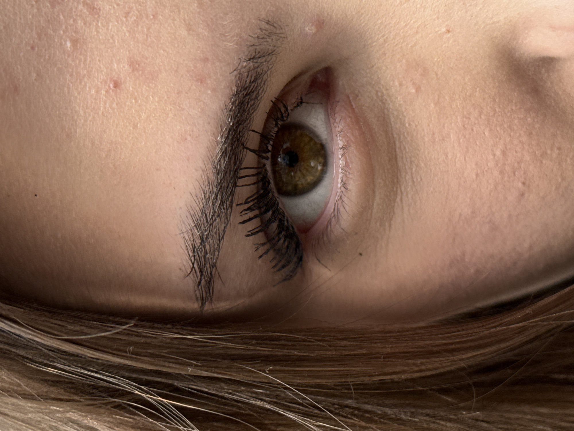 A close-up, slightly angled, downward shot of a person's face, predominantly featuring their right eye. The iris is a hazel color. The eyelashes are dark and appear to have mascara applied. A portion of the person's eyebrow is visible above the eye, and there is some skin texture suggesting the presence of minor skin imperfections near the eyebrow. The lower part of the frame includes strands of brown hair. The image does not contain sufficient information to infer the location in Sofia, Bulgaria or any activities.
