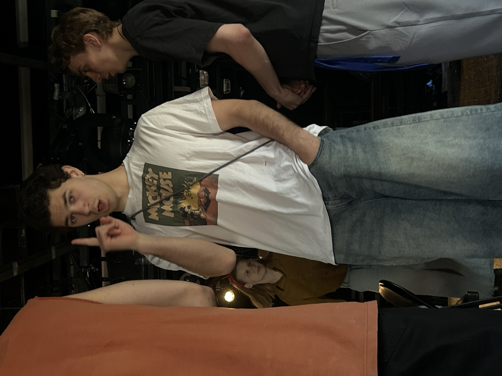 The image shows a group of four people in an indoor location that appears to be a theater backstage or a similar performance space.
The main focus is a young man in the foreground wearing a white t-shirt with a "Mickey Mouse" graphic, blue jeans, and a lanyard. He is pointing to the left with a surprised or sarcastic expression.
Above him, another young man in a black shirt and light-colored pants looks down, his hands clasped together.
Below the first man, a young woman in a brown shirt and a red headband looks upward toward a bright light. Her expression is not clearly visible.
Beneath her, a lower portion of a person wearing an orange shirt and black pants are also visible, with their arm extended forward.
The background is dark and cluttered, with various pieces of equipment.