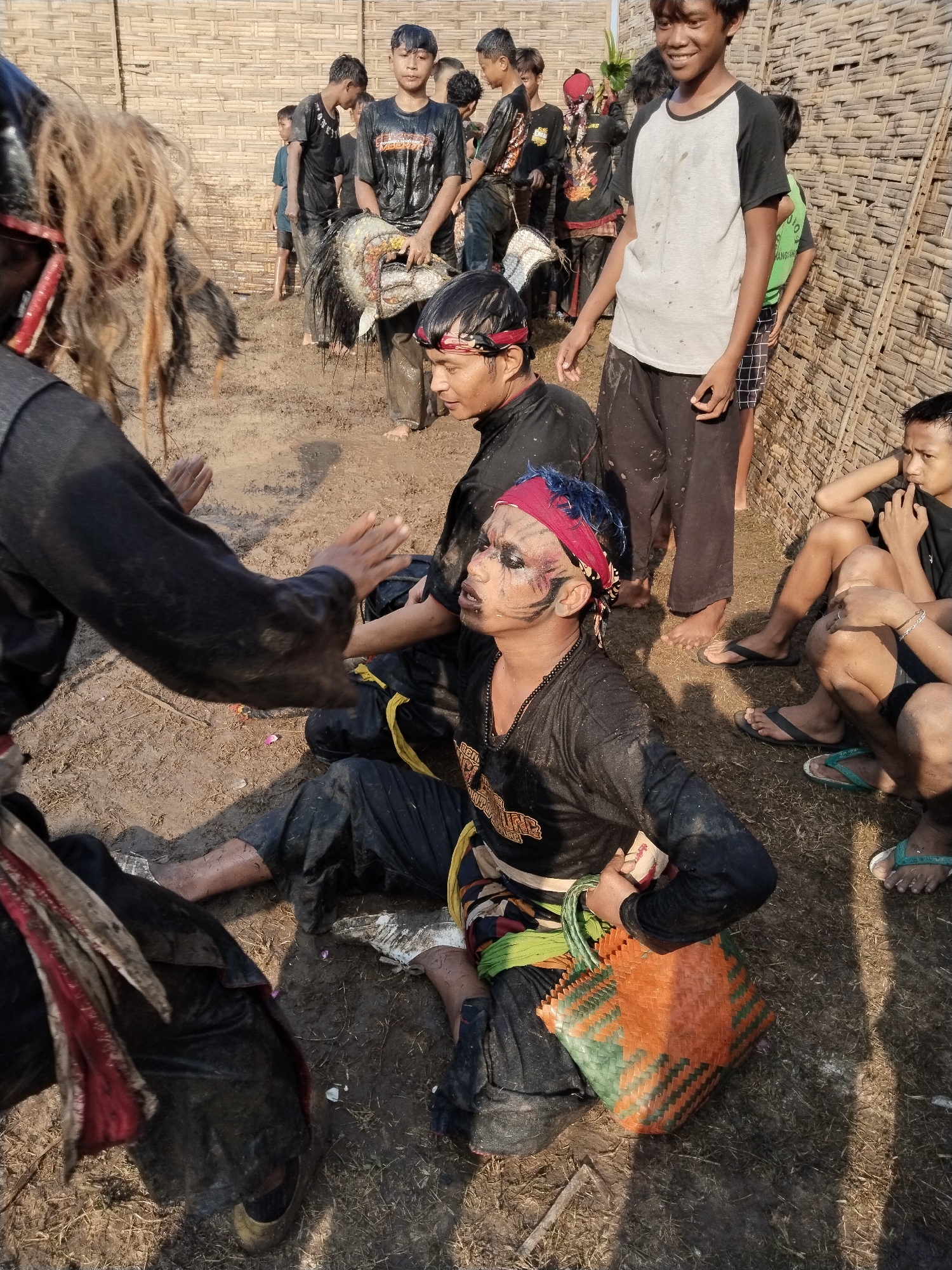 This image captures a group of young men and boys outdoors in Kedawung, Indonesia, seemingly during a break or after a traditional performance. The ground is muddy, indicating a physically demanding activity involving dirt and water.
In the foreground, a man with long, dark, unkempt hair and black clothing is partially visible on the left. His arm is extended towards a central figure, a young man seated on the muddy ground. This central figure, likely a performer, wears a black long-sleeved shirt, black pants, and a red bandana with streaks of blue hair. His face is elaborately painted with black, white, and red, resembling a tiger or mythical creature. He holds a brightly woven orange and green plastic basket. His expression suggests weariness or a state of trance.
Around these central figures, several other young men and boys are present. One boy in a white and black t-shirt stands smiling broadly in the background. Another boy leans against a woven bamboo wall, wearing a green sleeveless shirt and checkered shorts. Several others are holding traditional masks or headdresses, also covered in mud, suggesting they are props from the performance. These masks often represent mythical animals and are characteristic of Indonesian traditional trance dances such as Kuda Lumping or Jathilan.
The setting is outdoors, with a distinct woven bamboo wall forming the backdrop, a common feature in rural Indonesian environments. The bright daylight and visible shadows indicate it is daytime, likely late morning or afternoon. The participants are largely covered in mud, highlighting the immersive and physical nature of the event. While some appear tired, others seem relaxed and observing. Visible text on the face-painted man's shirt includes partial words like "GEMB" and "UNJ", and on a green shirt, "HANG ANJ". The scene offers a raw, authentic glimpse into a local cultural tradition.