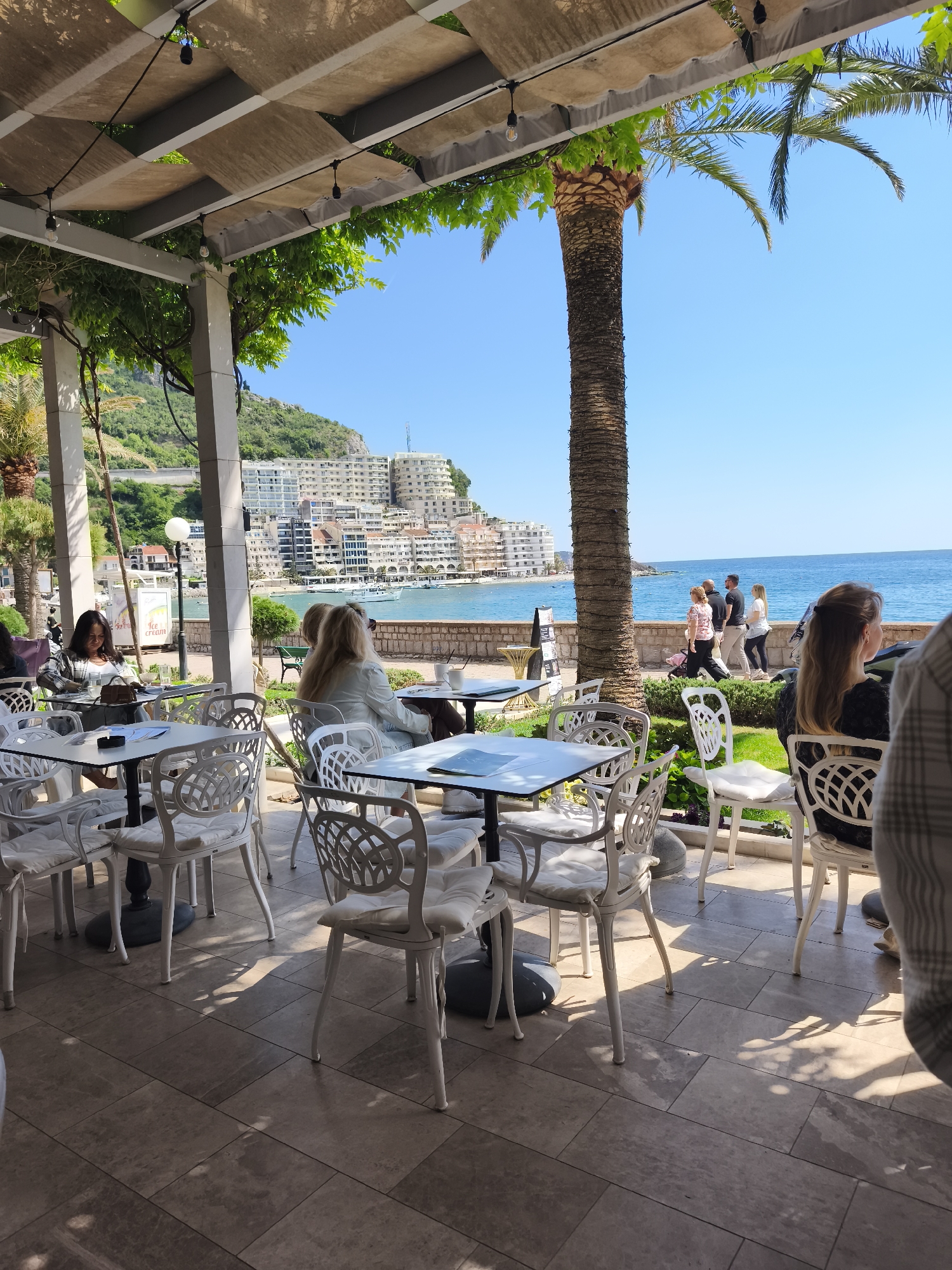 The image shows an outdoor seating area, likely a cafe or restaurant, in Rafailovići, Montenegro. Several white metal tables with chairs are arranged on a paved terrace. Diners are seated at some of the tables, including women in the foreground and left side of the image. In the background, a row of buildings lines the coast along the Adriatic Sea. A palm tree stands between the seating area and the coastal promenade where people are walking. An overhead pergola structure covers part of the seating area, decorated with plants. The weather appears sunny and clear.