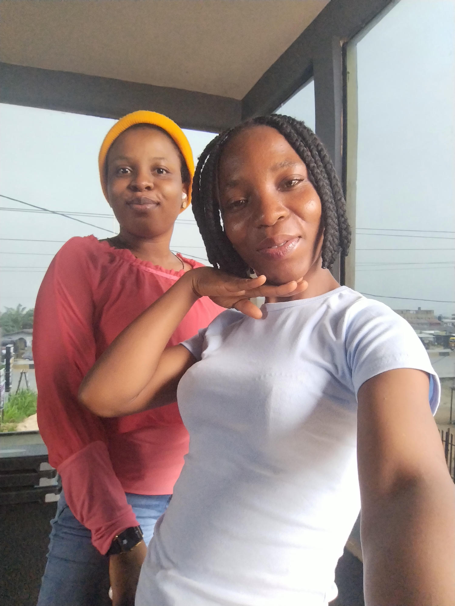 Two women are posing for a selfie outdoors in Uselu, Nigeria. The woman in the foreground is wearing a light blue t-shirt and has dark braided hair. She is resting her chin on her hand. The woman in the background is wearing a red long-sleeved shirt and a yellow beanie. She is standing behind and to the left of the woman in the foreground. They are standing on a balcony or porch with a railing. In the background, there are buildings and vegetation visible. The sky is overcast.