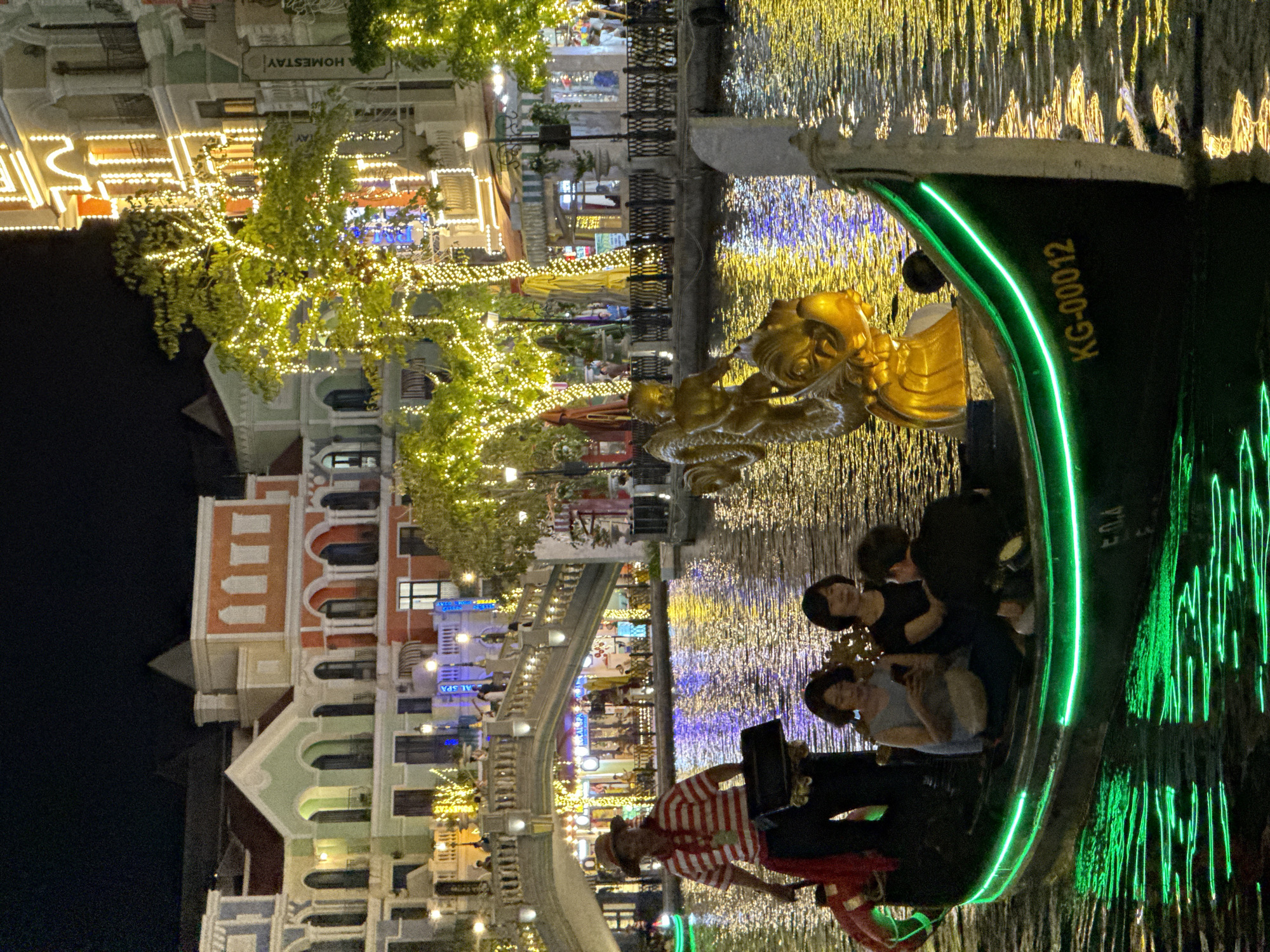 This night-time image captures a vibrant canal scene in what appears to be a themed resort area, likely Grand World in Phú Quốc, Vietnam. A dark-colored boat, resembling a gondola, occupies the foreground on the right, adorned with a bright green LED strip along its side. On the front of the boat is an elaborate golden sculpture, possibly a dragon or mythical creature. Text "KG-00012" is visible on the boat's hull.
Inside the boat, a group of people, including at least three passengers and a boatman, are enjoying a ride. The boatman, wearing a red and white striped shirt and a hat, is positioned at the front, possibly operating the vessel or performing. The passengers, primarily women, are seated and looking out at the illuminated surroundings.
The canal water glitters with countless reflections of warm-toned lights, creating a shimmering effect. On either side of the canal, multi-story buildings with varied architectural styles and colors line the banks. These structures feature arches, balconies, and intricate details, characteristic of Venetian-inspired architecture. Many of the buildings are brightly lit with string lights and other decorative illumination. Trees along the canal are also heavily decorated with small, golden string lights, enhancing the festive atmosphere.
A bridge arches over the canal in the mid-ground, also adorned with lights, with more distant buildings and streetlights visible beyond. In the upper left corner, the word "HOMESTAY" is partially visible on a building sign, and "AL SPA" can be seen on another building in the background. The overall scene suggests a lively and enchanting evening leisure activity, likely a scenic boat tour, under a clear night sky.