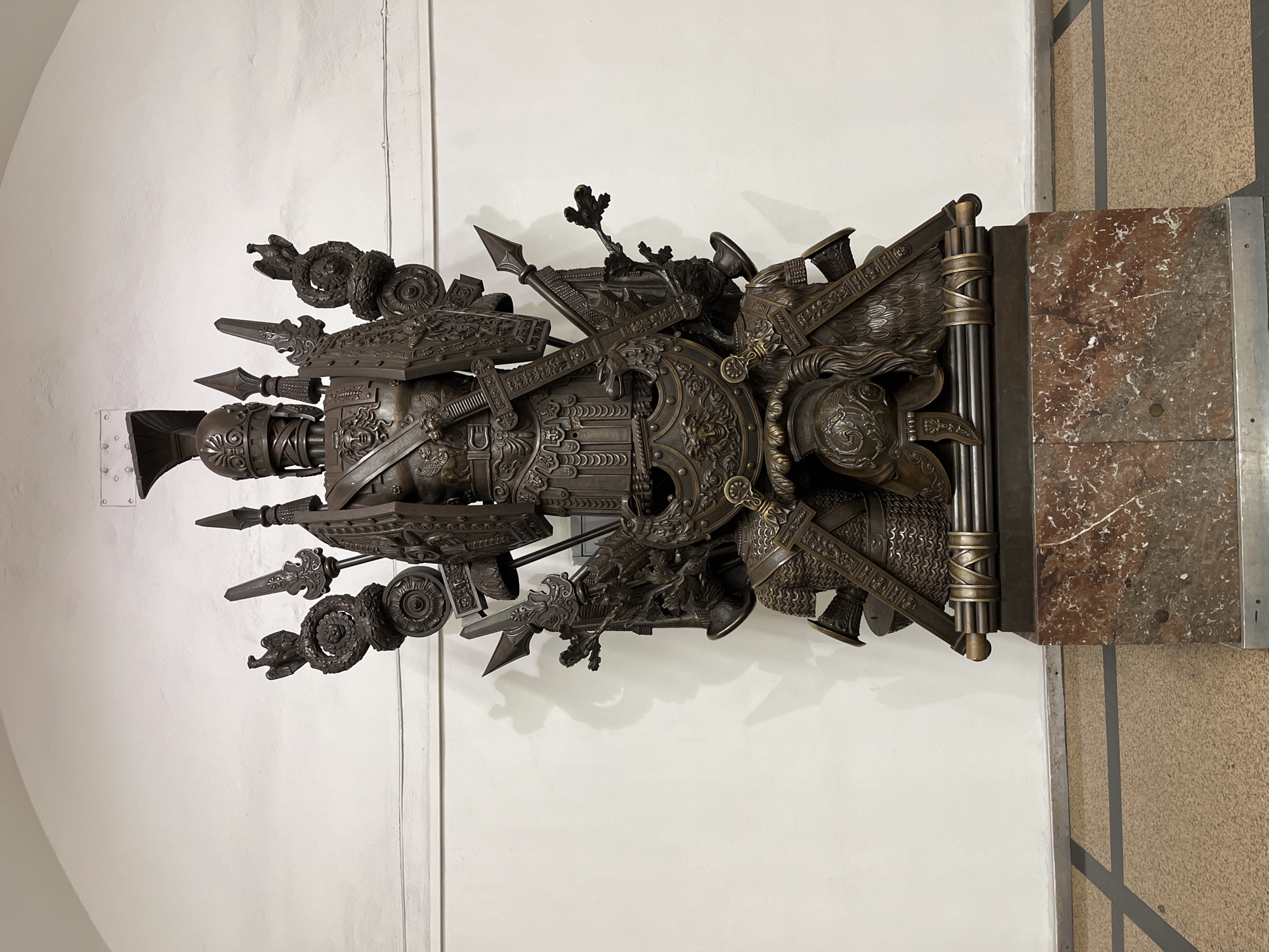 This image captures a highly detailed, ornate bronze sculpture against a stark white wall. The sculpture is a complex arrangement of military accoutrements and armor, suggesting a theme of warfare or historical commemoration. Prominent elements include shields adorned with intricate patterns, crossed swords, spears, helmets, and what appear to be laurels or wreaths. A central, helmeted figure's bust is partially visible, giving a sense of human presence within the collection of martial objects. The sculpture is mounted on a dark, possibly metallic, structure that extends to a base of polished red and white marble, with a textured wall of the same material visible to the right. The lighting is even, indicating an indoor setting, likely a museum or historical building in Saint Petersburg, Russia, given the location context. There is no discernible text in the image. The overall impression is one of grandeur, power, and historical significance.