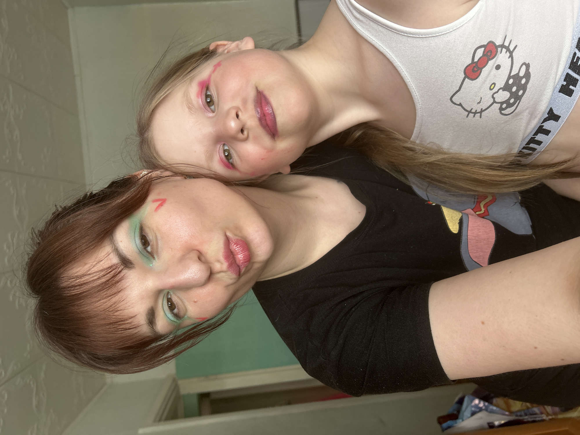 This image captures an indoor selfie of two females, an adult and a child, in a residential setting within the city of Фоки, Russia.
The adult female, with brown hair and bangs, is positioned on the left. She has green eyeshadow applied to her upper and lower eyelids and two small red 'V' shapes painted below the outer corner of each eye. Her lips are made up with red lipstick and puckered. She wears a black short-sleeved t-shirt with a partially visible colorful cartoon graphic on the front (a grey-blue form with pink/red and yellow accents). Her visible right arm exhibits fair skin with small brown macules.
The child female, approximately 6-10 years old, is positioned on the right, slightly in front of the adult. She has long, light-colored hair falling over her left shoulder. She wears bright pink eyeshadow on her upper eyelids, with two pink lines extending from the outer corner of each eye. Her lips are made up with a dark pink/purple glossy lipstick. She is wearing a white sleeveless crop top featuring a black and white "Hello Kitty" graphic with a red bow on the chest, and an elastic band below the bust reading "ITTY HE."
Both individuals are looking directly at the camera. The adult's head is tilted slightly, and the child's head is leaning into the adult.
The background consists of a light-colored ceiling with a textured, embossed pattern. To the immediate left, a doorway reveals an interior room with teal-colored walls. The wall directly behind the subjects is a plain, light beige or off-white color. The lighting is bright and even.