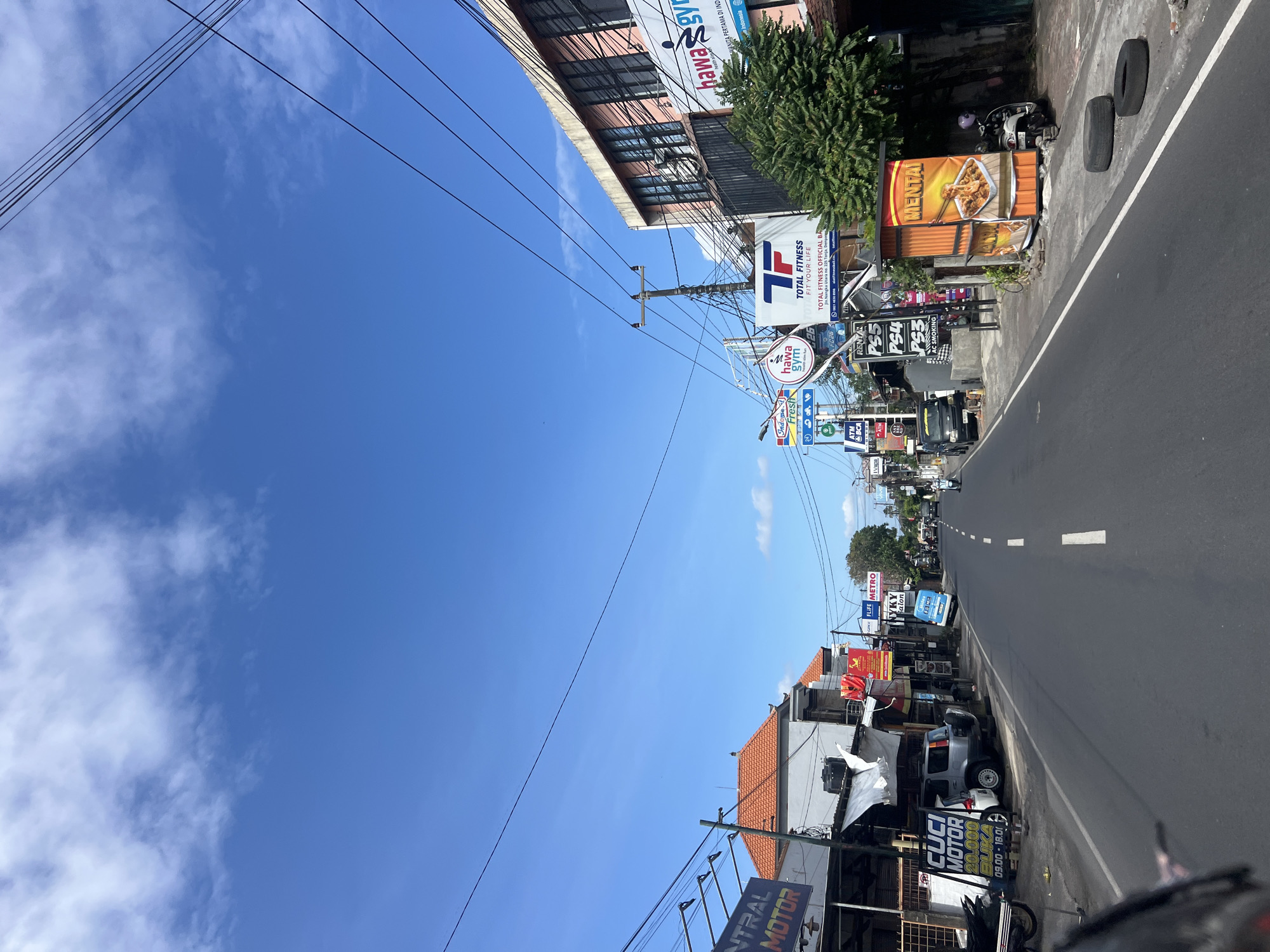A street-level, forward-facing view captures a bright, sunny day in Denpasar, Indonesia, with a clear blue sky and scattered clouds. The street is lined with buildings and storefronts displaying various signs for businesses such as a motor shop, a gym ("Hawa Gym" and "Total Fitness"), and a food vendor. A silver SUV and several motorcycles are parked along the roadside, and a few people are visible on the street, including a person riding a motorcycle in the distance. Overhead power lines crisscross the sky, and a few trees add greenery to the scene. The road is paved and has white dashed lines marking the lanes. The overall impression is of a typical commercial street in a bustling Indonesian city.
