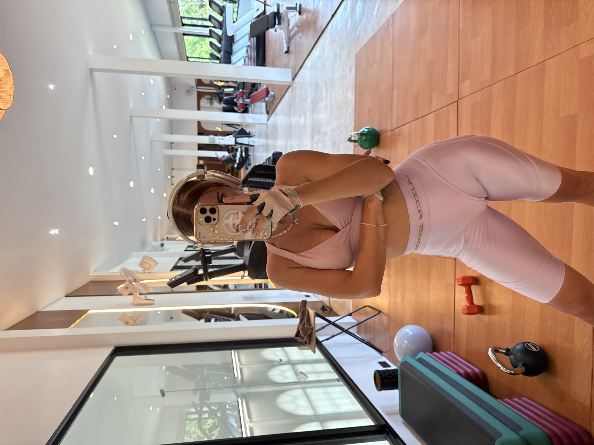 A female subject is captured in a mirror selfie within a brightly lit, spacious fitness center located in ตำบลเกาะพะงัน, Thailand. The subject, positioned centrally, is wearing a light pink athletic bra with a v-neck and wrap-around design, paired with light pink, mid-thigh length cycling shorts. The mirrored text "COOSƎ" (likely "E200C" or similar, reflected) is visible on the elastic waistband of her shorts. She wears beige fingerless workout gloves on her left hand, a white beaded necklace with a silver crescent moon pendant, and a thin white beaded bracelet on her right wrist. Gold over-ear headphones cover her ears, partially obscuring her brown hair. Her left hand holds an iPhone with a decorative, clear case, obscuring the upper portion of her face, while her right arm is bent and rests across her midsection.
The gym features a light brown wooden floor with visible seams. In the foreground, near the subject's feet, are a small black kettlebell with a silver handle, a small orange dumbbell, and a stack of teal and burgundy exercise steps. A white exercise ball and a black foam roller are visible further left near a large, black-framed window that reveals lush green foliage and distant arched window designs outside.
The background displays a modern gym environment with white pillars and a white ceiling fitted with numerous recessed spotlights. Mirrored wall sections alternate with vertical wooden panels featuring illuminated strip lights. Various pieces of exercise equipment are present, including multiple treadmills lined against a far windowed wall, weight benches with red pads, and other cardio machines. A small folding table with a brown towel rests near the left window. A woven, circular ceiling light fixture and a white ceiling fan are also visible in reflections.