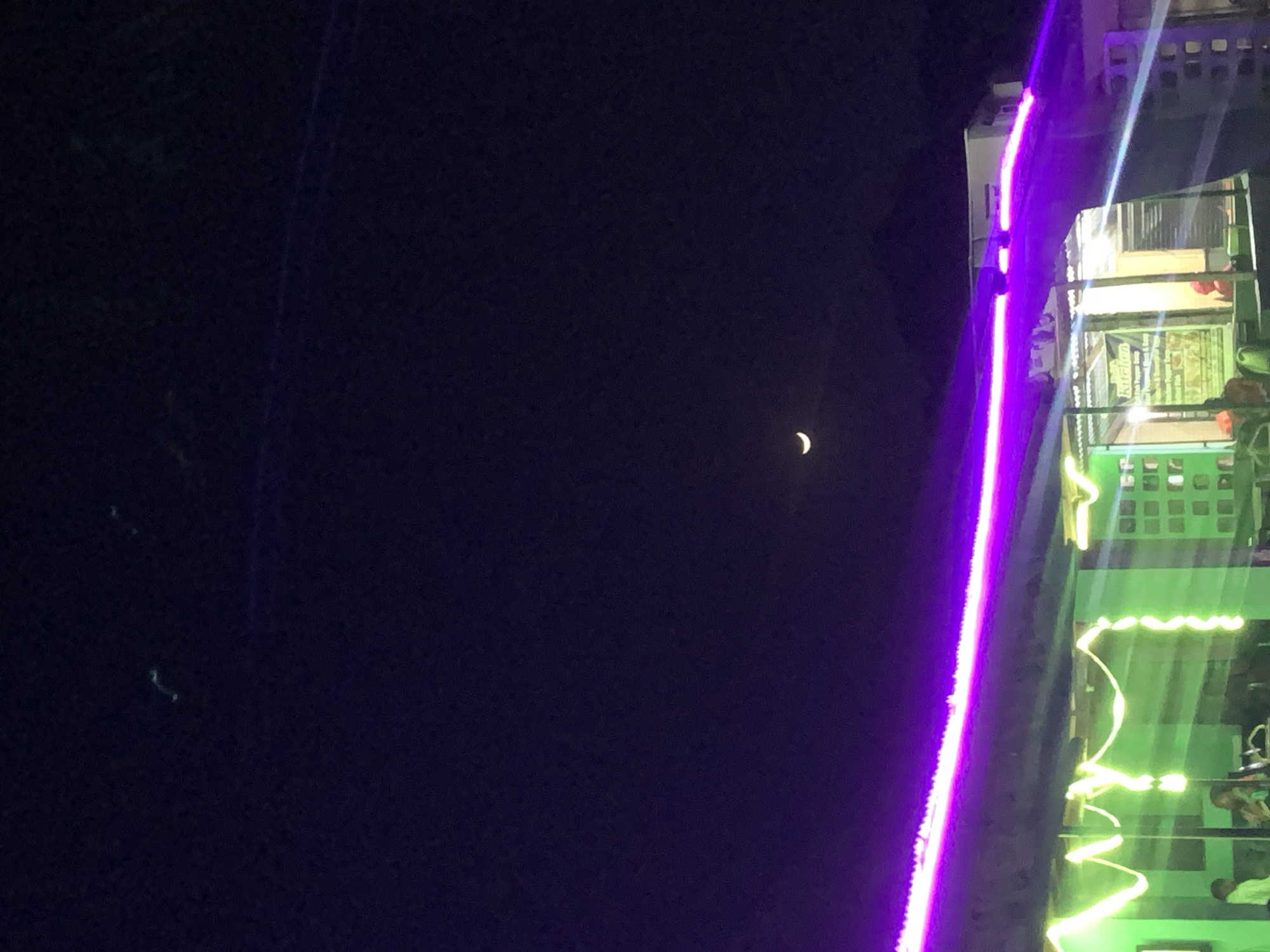 At night in New Stadium, Nigeria, a crescent moon is visible in the upper right quadrant of the frame. The majority of the frame is dark, with some indistinct shapes present. On the right of the frame is a building with green and purple lighting, with people inside and outside the building. The image is rotated 90 degrees.