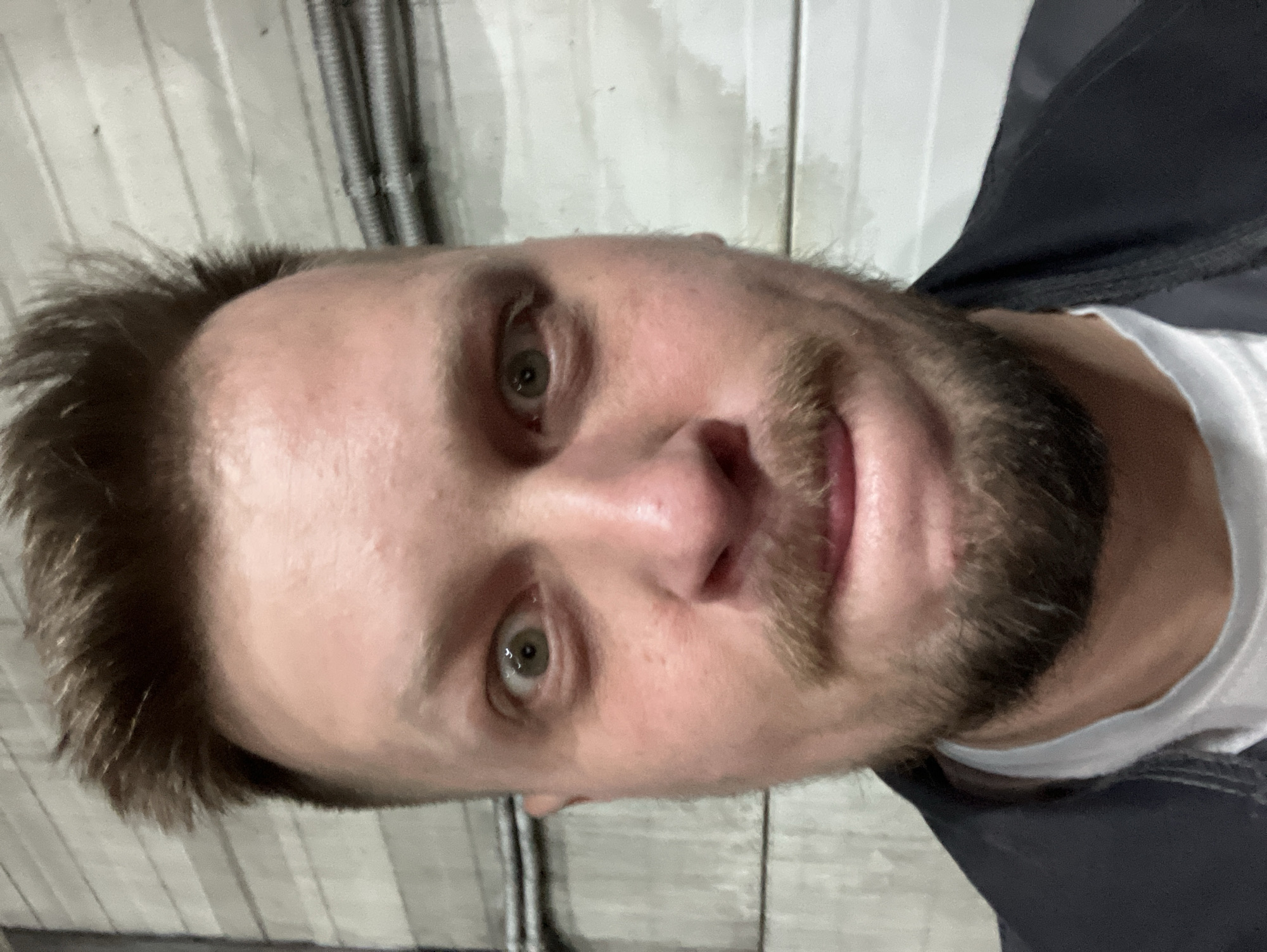 A close-up selfie of a fair-skinned male with brown hair and a beard. He is wearing a white t-shirt under a dark jacket. The background is a gray wall with vertical lines and some ribbed tubing. There is no indication of the city of Noviy, Russia in the image.