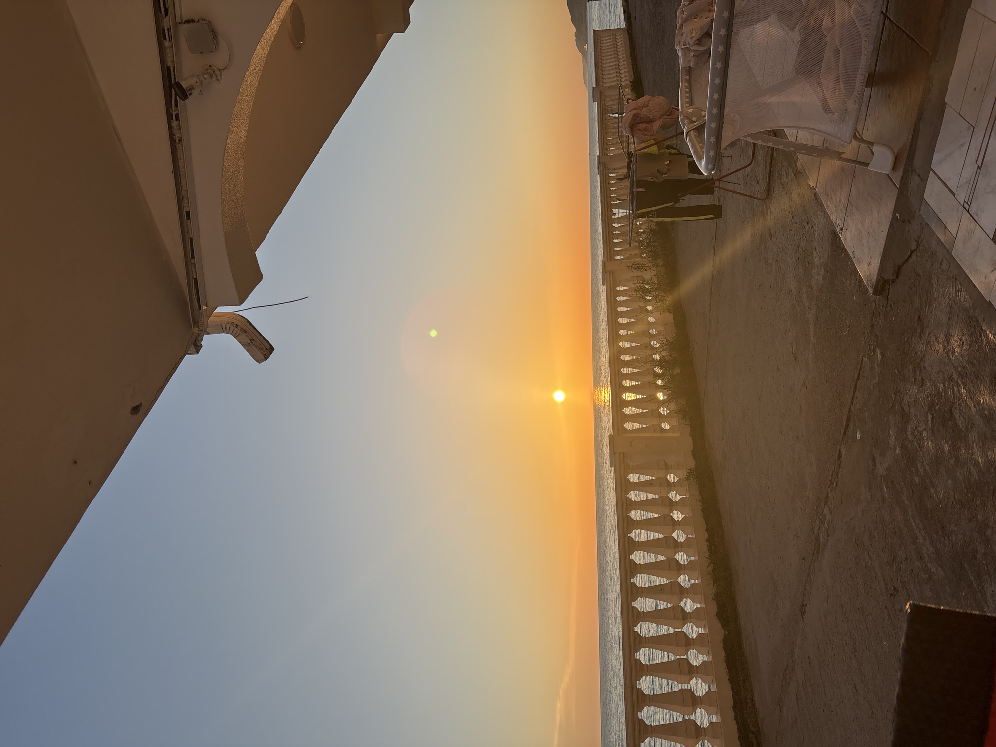 The image shows a seaside view at sunset. A building, likely part of a house or hotel, is visible on the left side of the frame. A white balustrade lines a patio or balcony overlooking the sea. There is a cot on the right. The sun is setting on the horizon, reflecting on the water. Given the context of Sochi, Russia, the location could be a coastal area near the city. No people are visible in the frame. The environment is calm and peaceful with a warm color palette.