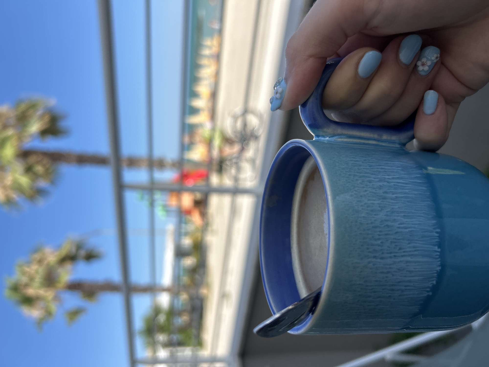 A close-up shot shows a person's hand, with light blue nail polish and delicate floral nail art, holding a textured, bright blue ceramic mug filled with coffee. A silver spoon rests inside the mug. The background is blurred, featuring a sunny outdoor scene with palm trees, a railing, and a beach with colorful umbrellas and lounge chairs visible in the distance. The overall impression is a relaxing moment enjoyed with a beverage in a seaside location, possibly Cupra Marittima, Italy, given the coastal scenery and palm trees.