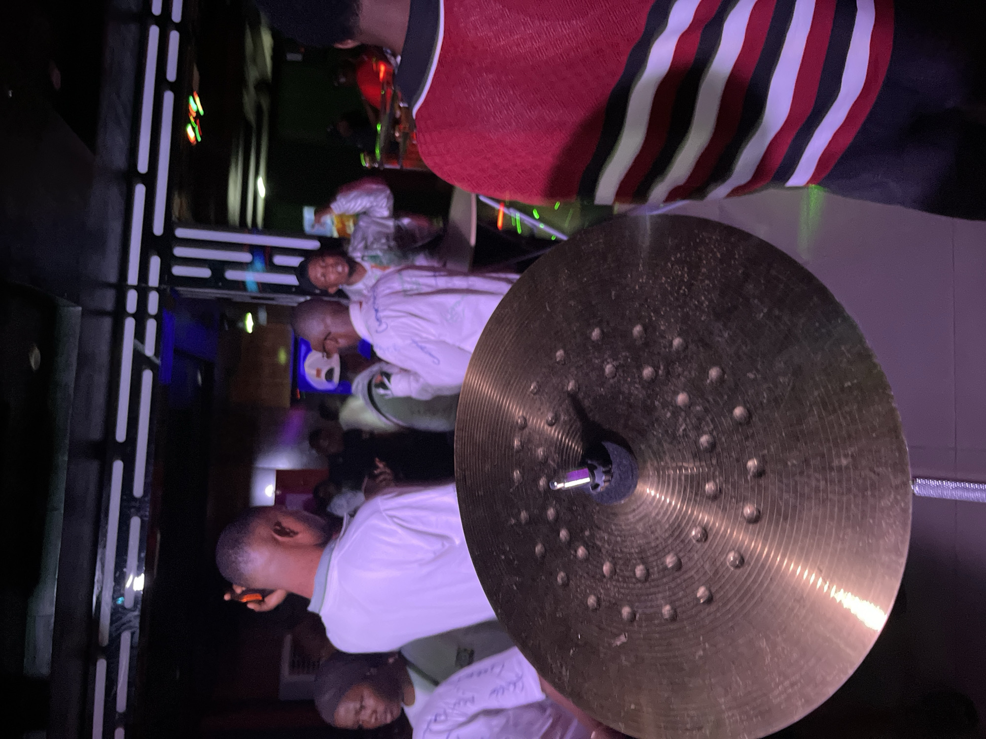 In an indoor venue, possibly a club or bar, a cymbal from a drum set is prominently featured in the foreground, angled towards the viewer. Behind it, a diverse group of individuals are visible, some engaged in conversation or holding drinks. The lighting appears to be dim and colorful, with a mix of purple and green hues illuminating the scene. A person wearing a red and black striped shirt stands on the right side of the frame, partially obscuring the background. The overall atmosphere suggests a social gathering or event. The location is described as Unknown, Unknown.