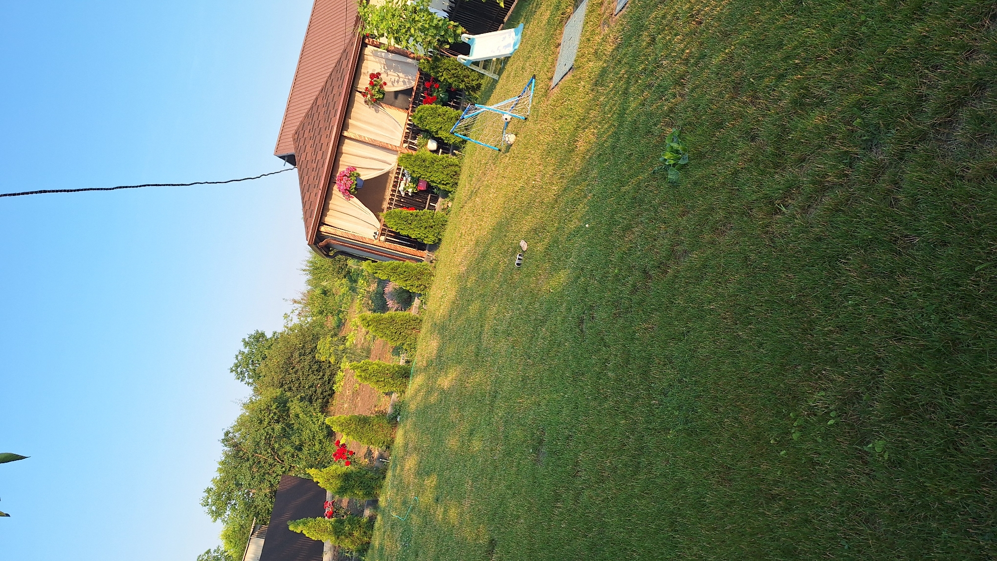 The image depicts a sunlit backyard in the city of Sărata-Galbenă, Moldova. A large expanse of manicured green grass dominates the foreground, sloping gently upwards towards a raised garden bed lined with small, conical evergreen trees and red flowers. In the middle ground, a brown-roofed gazebo with cream-colored curtains is visible, adorned with hanging flower baskets. Adjacent to the gazebo, a small blue and white soccer goal with a ball, and a white children's slide are set up on the lawn. A dark, thin cable hangs diagonally across the top right of the frame against a clear blue sky. Further back, a brown wooden fence separates the property from a wooded area with lush green trees. The overall scene suggests a well-maintained outdoor recreational space.
