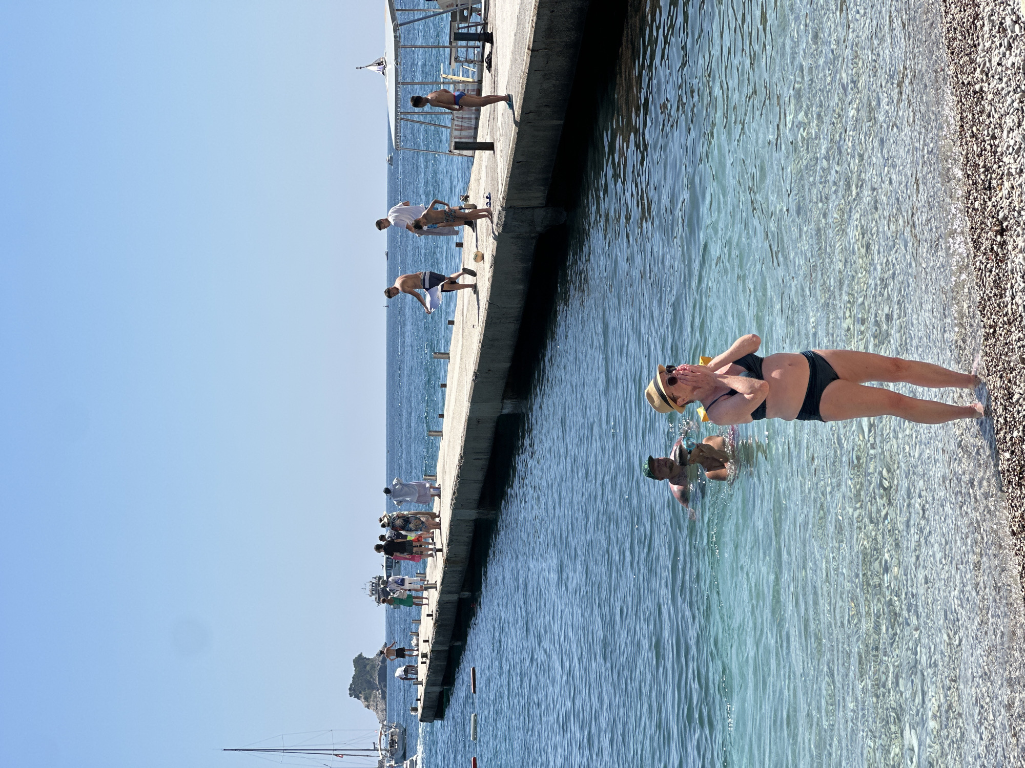 The media file depicts a bright, sunny day at a beach in Budva, Montenegro. The scene is a long, concrete pier extending into clear, blue water. Several people are on the pier, some wading in the shallow water at the edge, while others are standing or walking further down. A few individuals appear to be preparing to jump or dive into the water.
On the pier, a group of people are scattered along its length. Closer to the viewer, a woman in a dark bikini and a straw hat is standing on the pebbly shore, with her hands covering her face. Beside her, a person is in the water with a snorkel mask. Further down the pier, a young boy in swimming trunks stands near an adult in a white shirt and dark shorts. Another person in blue swim trunks stands near them.
The water is clear, revealing the pebbly bottom, and the sunlight reflects off its surface, creating a shimmering effect. The sky is a vast expanse of clear blue, indicating fair weather. In the distance, several boats are visible, including sailboats, and a small, rocky island with some vegetation can be seen offshore. The overall atmosphere is one of leisure and summer vacation. There is no visible text in the image.