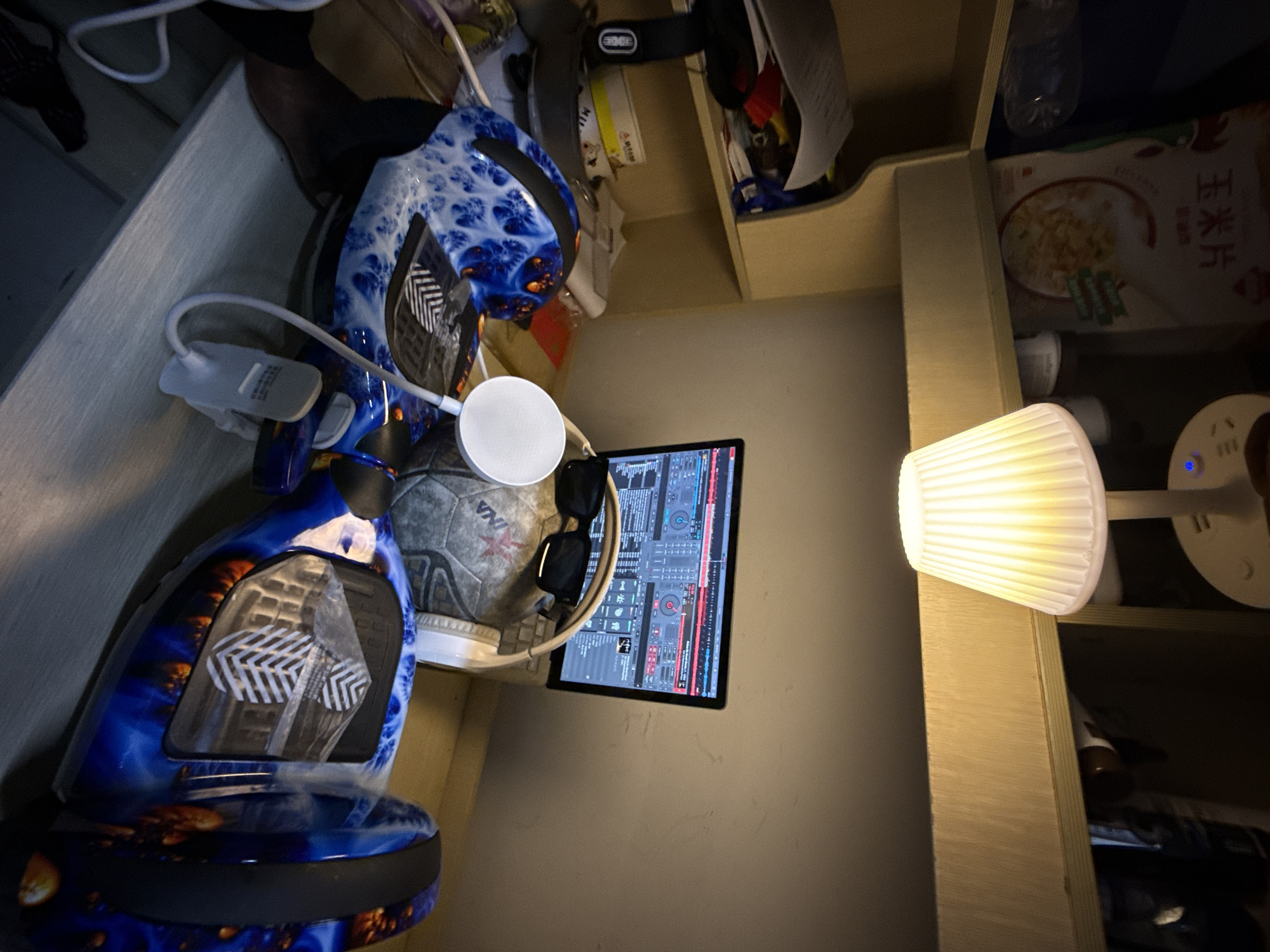 The image displays a cluttered indoor personal space, likely a desk or shelf unit, in dim lighting, suggesting nighttime. The primary subject in the foreground is a blue and orange patterned hoverboard or self-balancing scooter. A small, white clip-on light is attached to the hoverboard, and a white circular object, possibly a wireless charger or smart device, rests nearby.
Adjacent to these items is a black tablet or laptop, displaying a complex interface of DJ software with various waveforms, faders, and controls. A pair of black sunglasses is placed on the surface next to the tablet. A grey and white soccer ball, with the word "VIVA" visible, is also part of this arrangement.
The objects are situated on and around a light-colored wooden shelf unit. On one shelf, a white pleated desk lamp provides illumination, casting a warm glow. Another white circular device with a blue indicator light, possibly a smart speaker or multi-port charger, is visible to the right of the lamp. The background shows various items stored on the shelves, including boxes, bottles, and papers. A packet of cereal, clearly labeled "玉米片" (yùmǐpiàn - corn flakes) in Chinese characters, is visible on an upper shelf. In the darker background, a black strap or band has the word "EDGE" visible. The overall scene depicts a functional, albeit crowded, personal area in a room, consistent with a home or dormitory setting in Jiangjin, China.