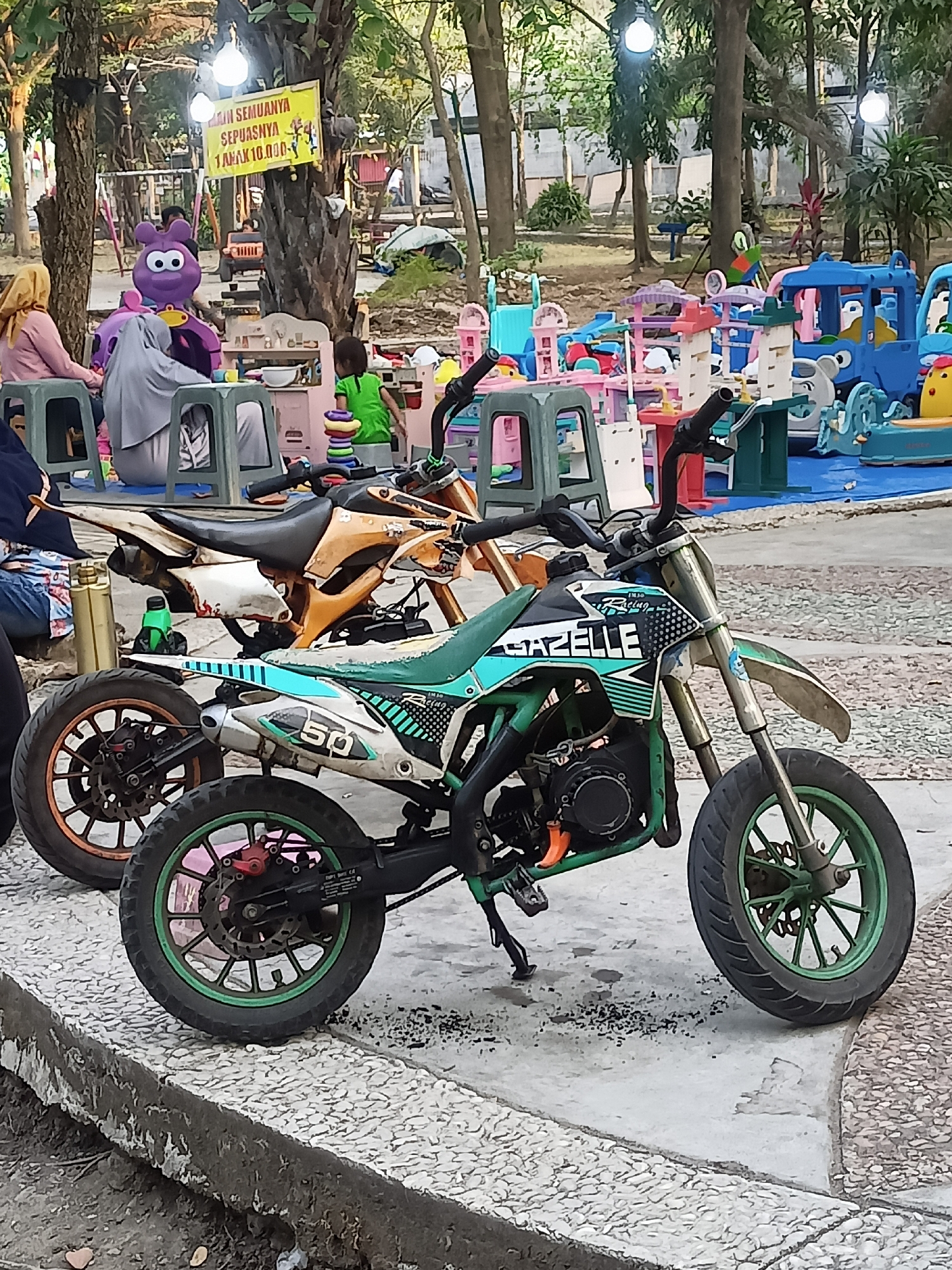 In Girimargo, Indonesia, two dirt bikes are parked on a textured concrete area. The foreground is dominated by a green and white dirt bike, clearly labeled "GAZELLE," with "50" visible on its side. Its wheels have green rims. Behind it, partially visible, is an orange and white dirt bike.
The background reveals a children's play area, suggesting a public park or amusement facility. Several colorful plastic toys and playground equipment are scattered around, including what appears to be a blue cartoon-like vehicle and various structures that could be playhouses or shops. In the mid-ground, two adults are seated on plastic chairs. One wears a light pink outfit and yellow headscarf, while the other is covered in a grey hijab. A large, purple, plush character with big eyes stands to the left of the seated individuals. A yellow sign with Indonesian text is attached to a tree trunk, indicating pricing for play activities. Another child, wearing a green shirt, is visible playing near the toys. The environment is outdoors, with trees and foliage indicating a park-like setting. Natural light suggests it is daytime.