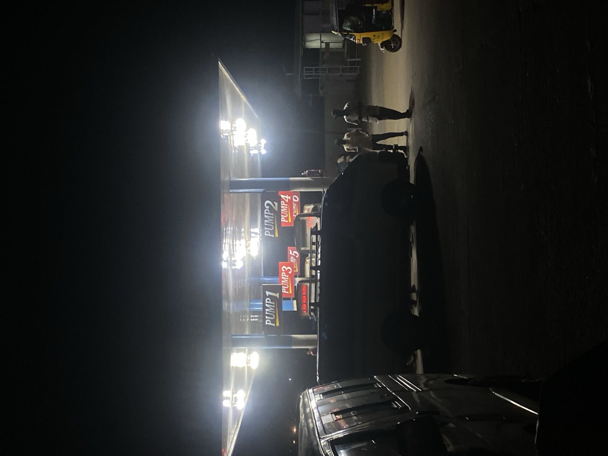 The image, rotated 90 degrees clockwise from its likely original orientation, captures a brightly lit gas station at night in Tudun Wada, Nigeria. Powerful overhead lights from the pump canopy intensely illuminate the immediate area, creating a stark contrast with the deep shadows and dark surroundings.
Several individuals are visible standing near the fuel pumps, mostly as silhouettes against the bright light, appearing to be engaged in activity or waiting. A dark-colored SUV or similar vehicle is positioned at one of the pumps, while another vehicle is partially visible in the bottom foreground. In the background, a distinct yellow and black three-wheeled auto-rickshaw is parked.
Visible text on the fuel dispensers clearly identifies them as "PUMP 1", "PUMP 2", "PUMP 3", "PUMP 4", and "PUMP 5". The scene portrays typical night-time operations at a petrol station, with vehicles being refueled. The strong artificial lighting at night, combined with the presence of the auto-rickshaw, provides clear temporal and regional cues.