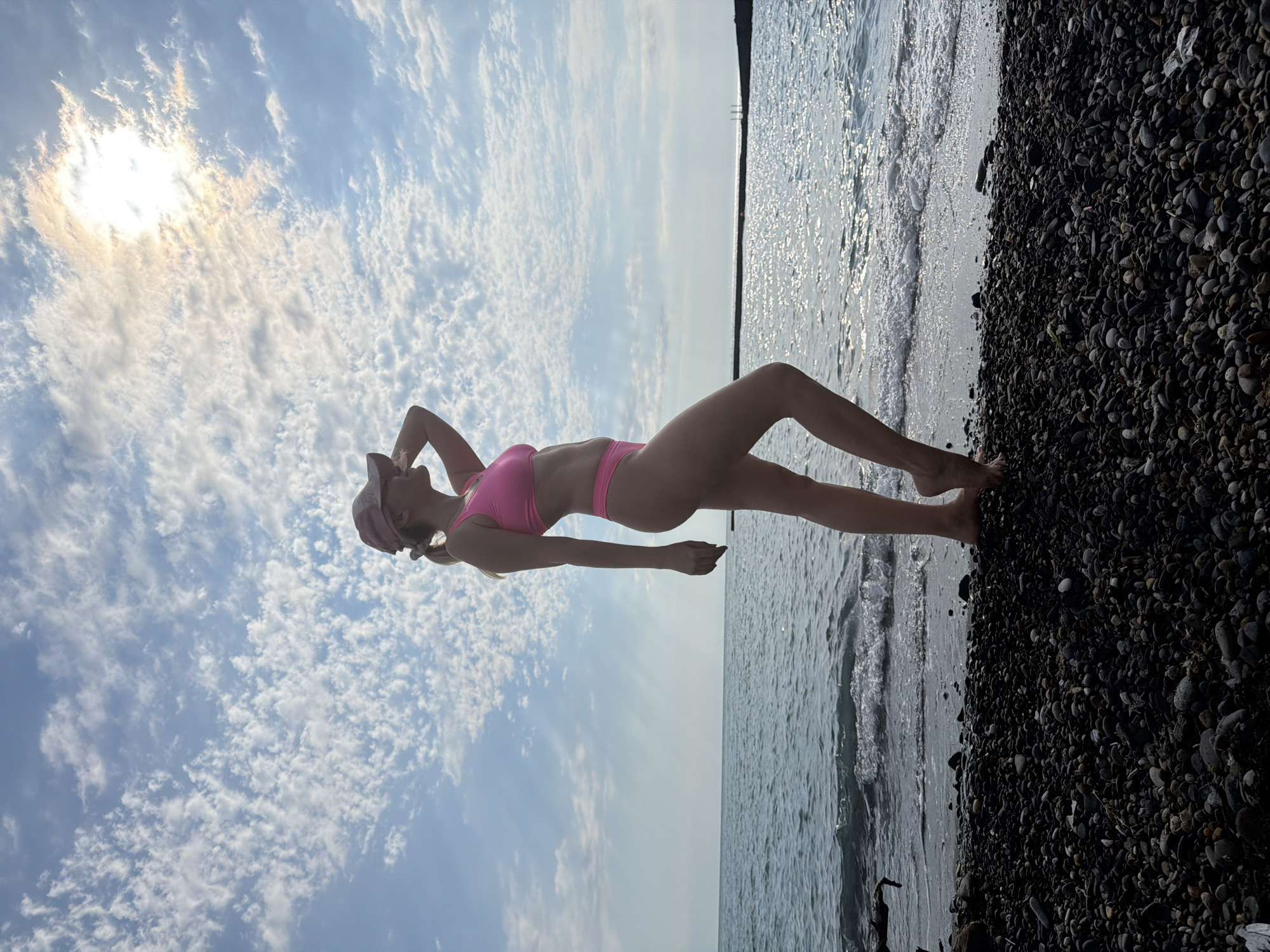 The image depicts a woman standing on a pebble beach, facing the sea. She is wearing a pink bikini and a pink baseball cap, with her right hand touching the brim of the cap. The sun is shining brightly in the sky, partially obscured by clouds, creating a shimmering effect on the water. The sea is calm with gentle waves lapping at the shore. The beach is covered in dark, smooth pebbles. In the distance, a man-made structure, possibly a pier or breakwater, is visible extending into the sea. The sky is a clear blue with scattered white clouds. The overall scene suggests a serene and sunny day at the beach in Sochi, Russia.