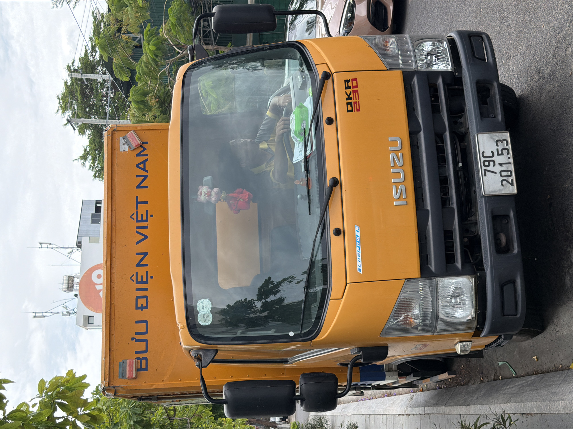 A ground-level, medium-shot, rotated perspective reveals an orange Isuzu delivery truck oriented towards the viewer in Bac Nha Trang Ward, Vietnam. The truck is positioned on asphalt, suggestive of a street or parking area. The license plate is visible, reading "79C 201.53." The back of the truck has blue lettering which, when rotated, reads "BƯU ĐIỆN VIỆT NAM". The driver is visible through the windshield, wearing a yellow and dark shirt. A building is in the background with the "VNPT" logo visible, suggesting a telecommunications building. Tree foliage frames the edges of the image.