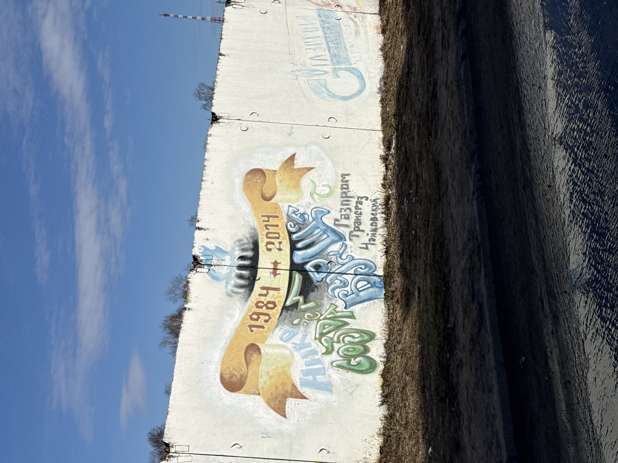 The image shows a concrete wall with graffiti. The graffiti includes the years "1984" and "2014" on a banner, and text in Cyrillic script, including the words "Газпром Трансгаз Чайковский" (Gazprom Transgaz Tchaikovsky). The wall appears to be near a body of water. The sky is blue with clouds in the background. The location is Unknown, Unknown.