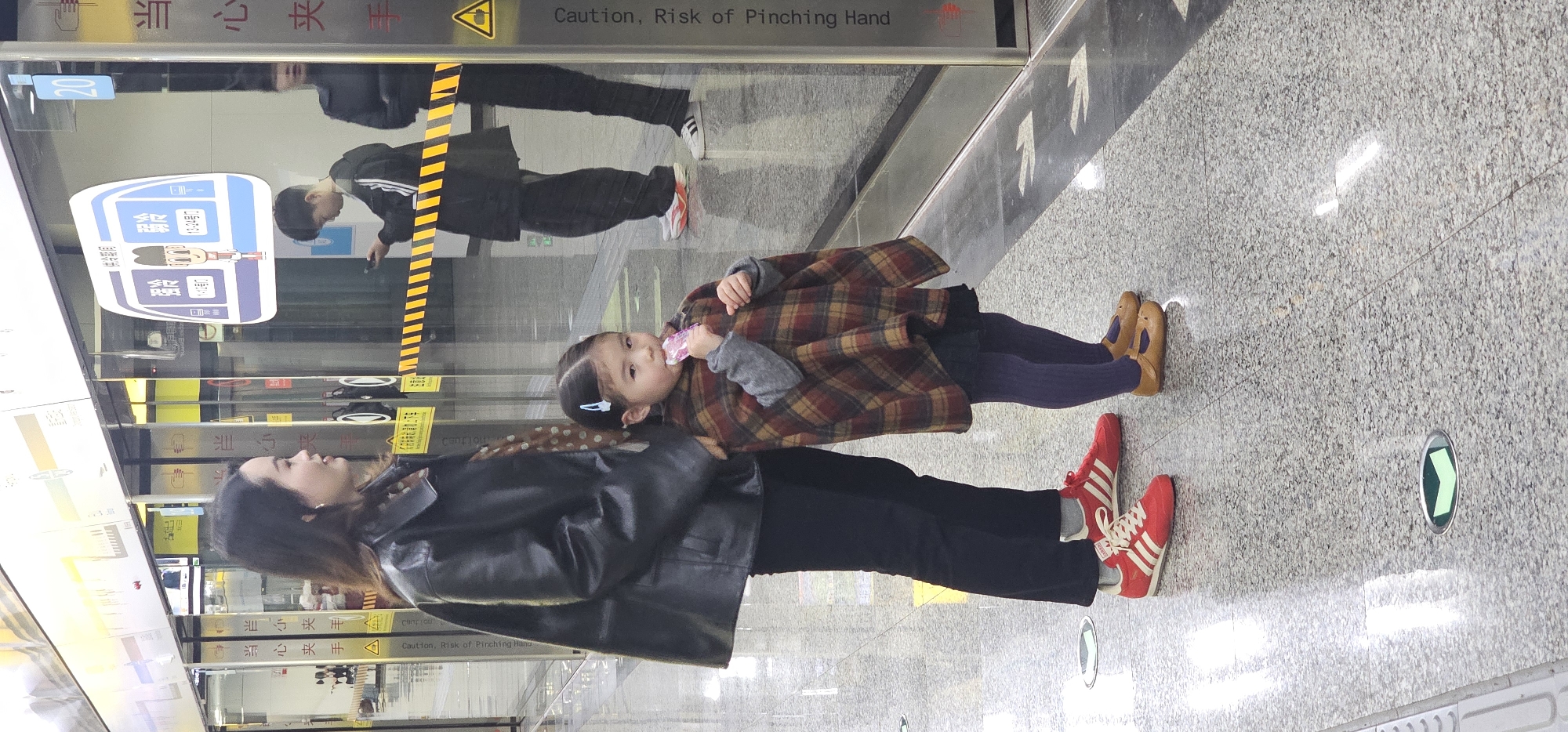 The image captures a scene at a metro station, potentially in the Jiulongpo District of China, as indicated by the Chinese characters on the glass screen. A woman wearing a black leather jacket and black pants stands next to a young girl with a hair clip in her hair, who is eating something. The girl is wearing a plaid poncho, navy leggings, and tan shoes. The woman is wearing red Adidas sneakers with white stripes. A glass screen separates them from the train platform. Reflected in the glass is another individual wearing a black tracksuit with white stripes. The platform floor is a speckled gray. Arrows are visible on the floor of the train platform. There are signs in Chinese on the glass. One sign has an illustration of a cartoon character. The English text "Caution, Risk of Pinching Hand" is on the screen.