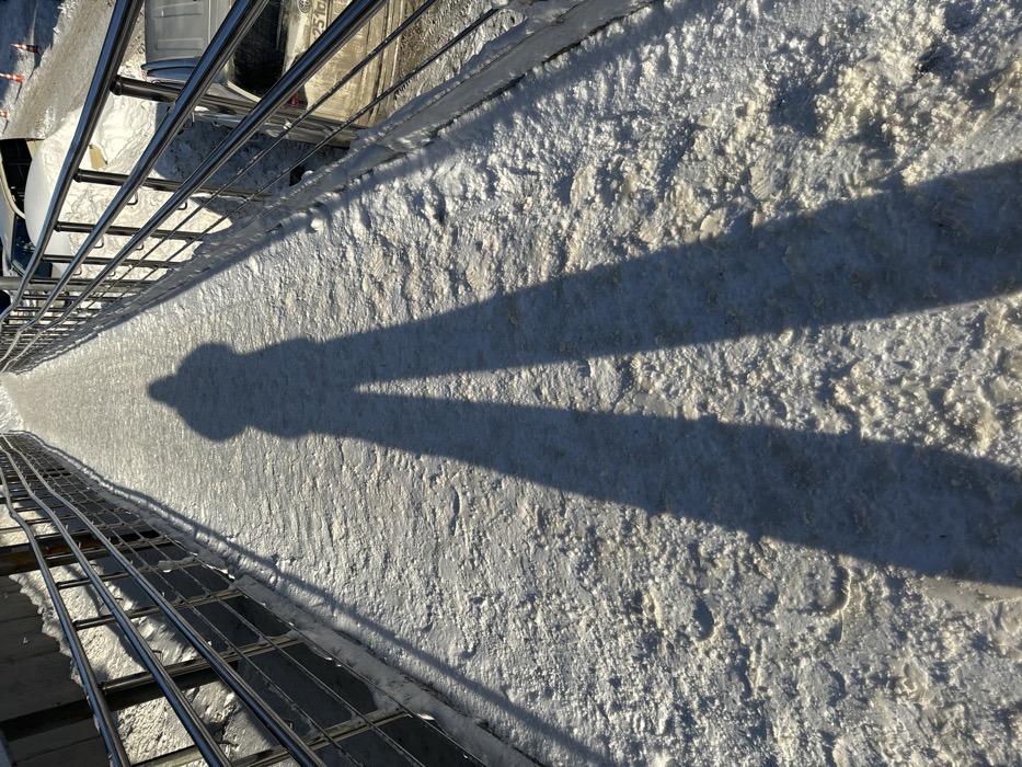The image depicts a snow-covered pedestrian ramp or bridge under clear, low-angle sunlight, casting a prominent shadow of a person. A long, distinct human shadow, likely belonging to the photographer and indicating a hooded figure, stretches from the bottom center of the frame along the snow-covered walkway towards the upper center. The walkway surface is uneven, consisting of compacted snow and ice, with visible textured patterns and faint footprints. Polished metal railings, appearing to be stainless steel with multiple horizontal bars, line both sides of the pathway, extending into the background. To the left of the walkway, beyond the railing, is a snow-covered embankment or raised concrete structure. To the right, at a lower elevation, a street or parking area is visible with several vehicles, including a light-colored sedan partially obscured by snow, and orange traffic cones in the distance. The overall environment suggests winter conditions in an urban or suburban setting. Specific street or landmark identification within Shul'gino, Russia, is not discernible from the visual information provided.