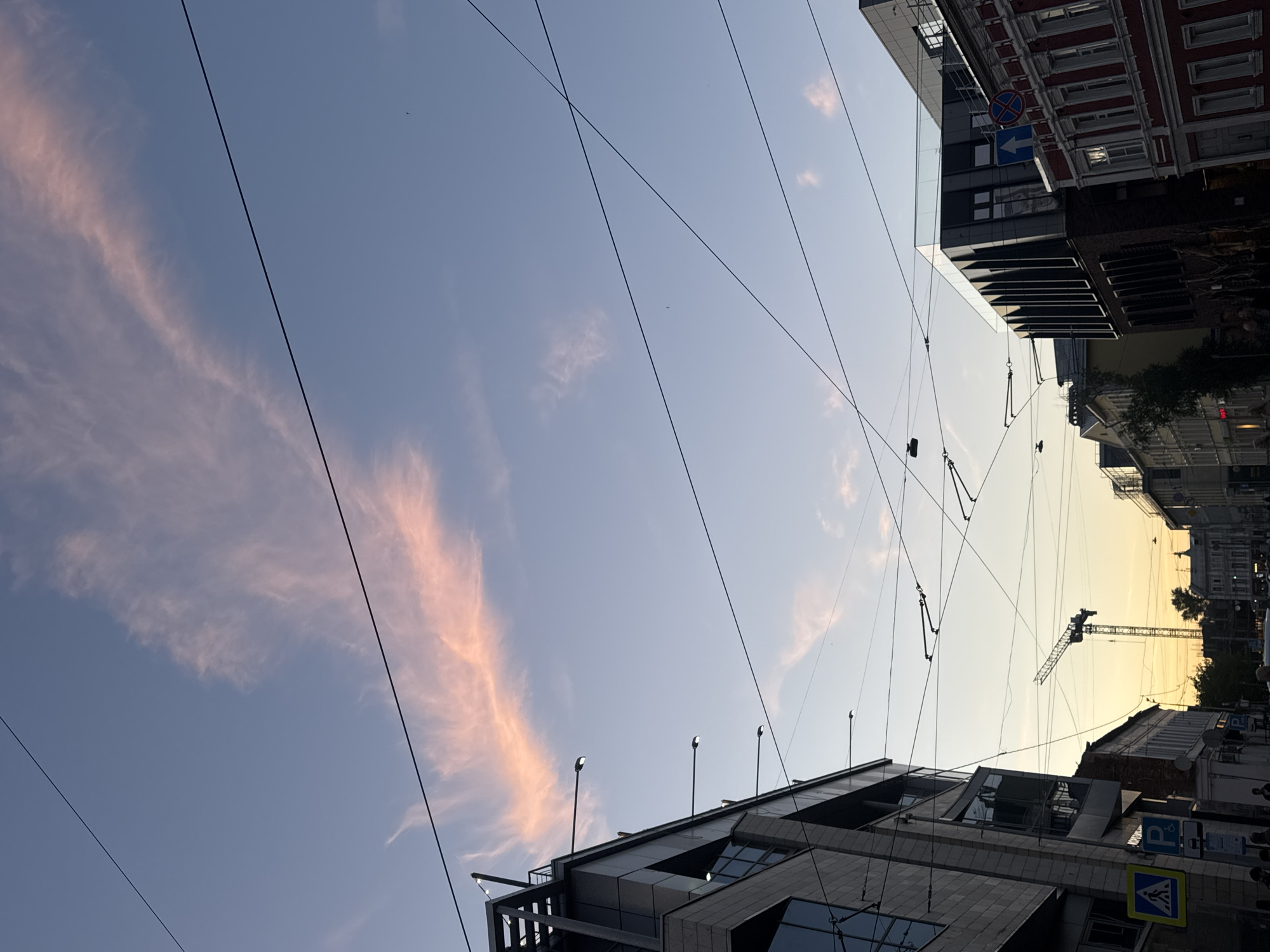 The image captures a street scene in Nizhny Novgorod, Russia, during what appears to be sunset or twilight. The sky is a gradient of soft blues and pinks, with wispy clouds illuminated by the setting sun. Overhead, a complex network of electrical wires, likely for trams or trolleybuses, crisscrosses the frame.
On the left, a modern, multi-story building with a glass facade and dark structural elements dominates the foreground. Streetlights extend from its side. To the right of this building, a construction crane stands tall in the middle distance, silhouetted against the warmer hues of the sunset sky.
Further down the street, various other buildings are visible, including a red-brick structure with traditional architectural details on the far right, and other edifices with varying architectural styles and colors. Pedestrians can be seen in the lower part of the image, though they are not the primary focus. Street signs, including a pedestrian crossing sign, are also present. The overall impression is a contemporary urban environment transitioning into evening.