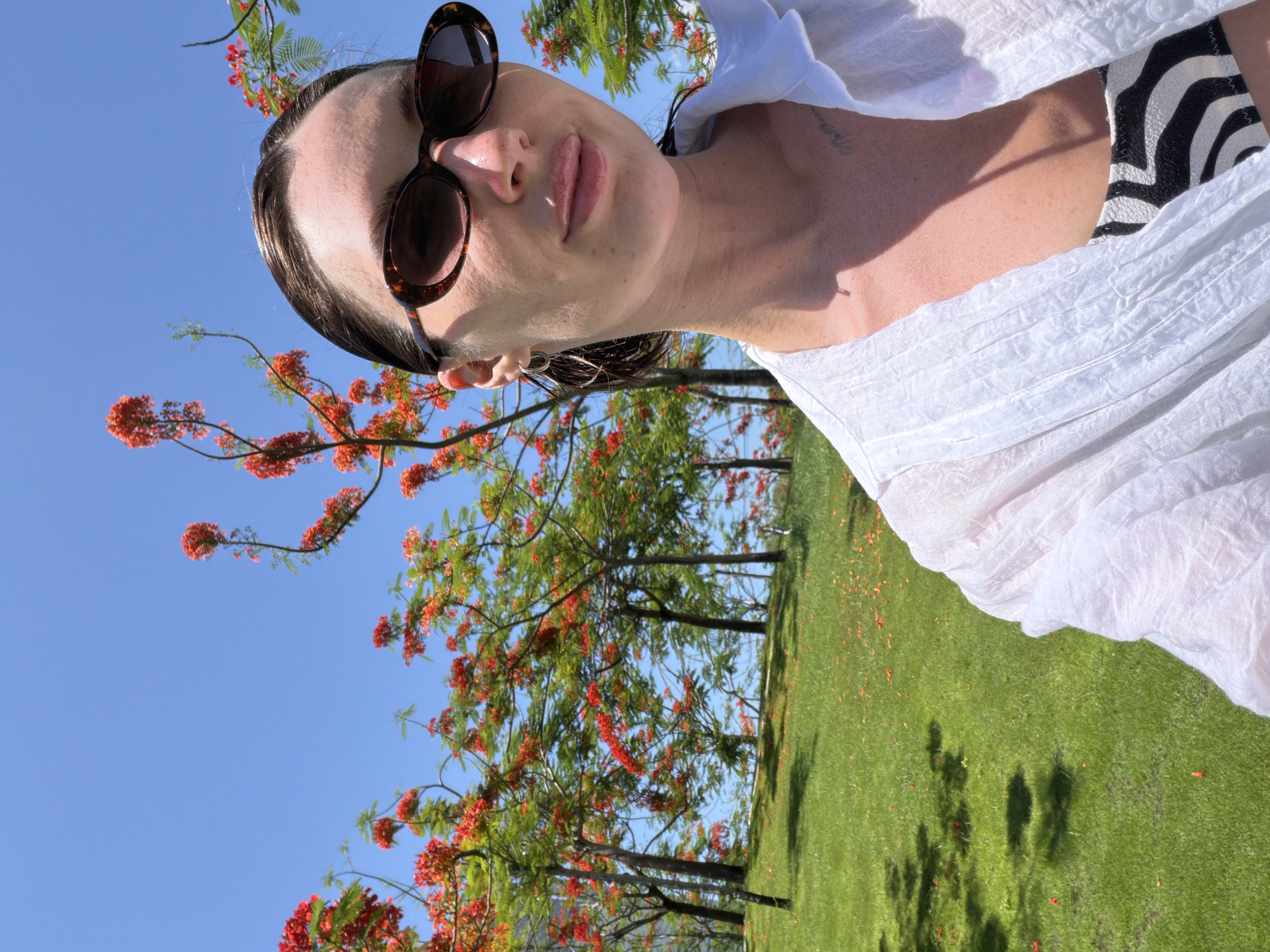 A medium shot captures a woman outdoors in what appears to be a park or garden setting, with a clear blue sky and a row of trees with vibrant red flowers in the background. The woman is facing the camera, with her right side closer to the viewer. She has dark hair pulled back and is wearing tortoiseshell sunglasses and a white, loosely woven shirt over a black and white striped top. Her expression is neutral, and her lips are slightly parted. To the left of the woman, the scene transitions to a grassy incline dotted with fallen red petals. Shadows of tree branches are cast on the grass. The overall atmosphere suggests a sunny, warm day. The location is identified as Dong Ninh Hoa Ward, Vietnam.