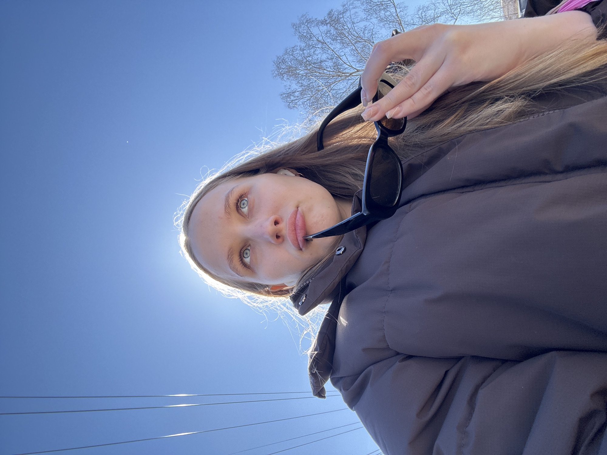 A young woman with fair skin and light brown hair is taking a selfie on a sunny day in what appears to be Chaikovsky, Russia. She is wearing a dark puffer jacket and holding a pair of sunglasses. The background includes a clear blue sky, power lines, and a tree with bare branches. The sun is shining brightly, causing a glare around her head.
