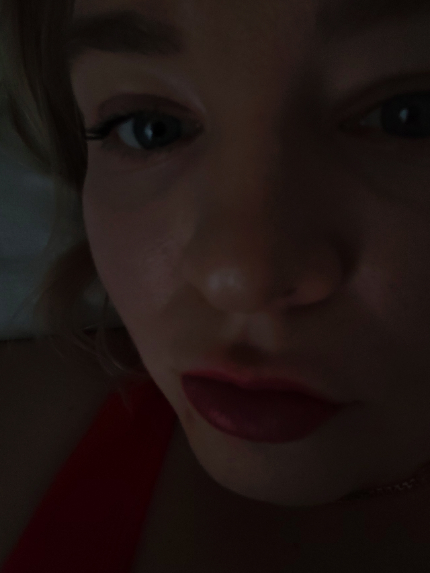 A close-up shot of a fair-skinned person with blue eyes and red lipstick. The lighting is dim, obscuring some details. The person is wearing a red top, and a necklace is visible. The location is not discernible.