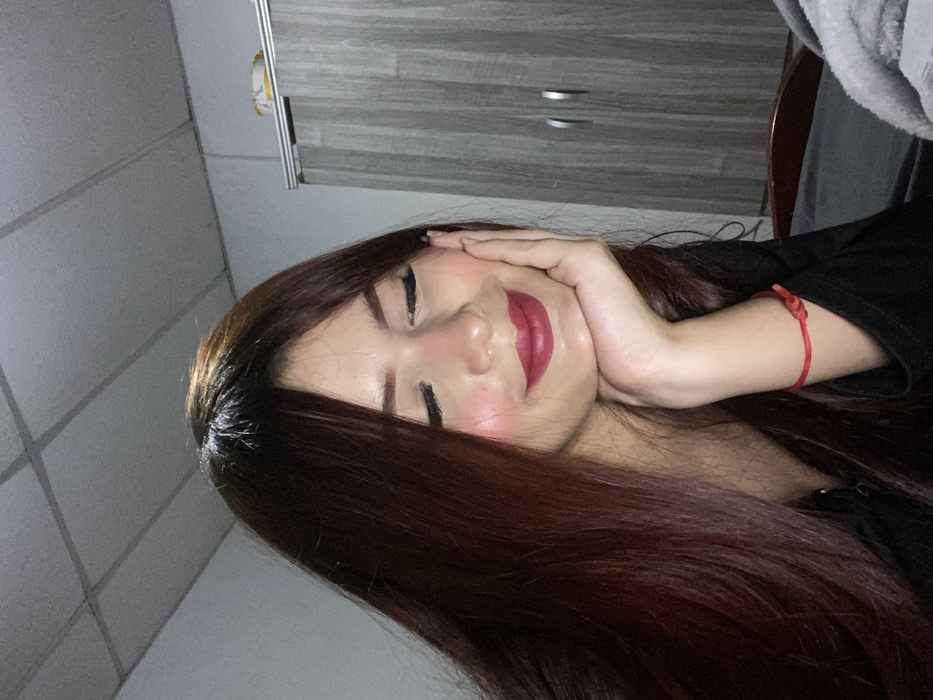 A woman with long, straight brown hair poses with her hand against her cheek. Her eyes are closed and she wears dark lipstick. She has a red string tied around her wrist. The background includes a gray cabinet with silver handles, a light-colored wall with paneling, and a gray fabric drape. The photo is taken from a slightly sideways, low angle.