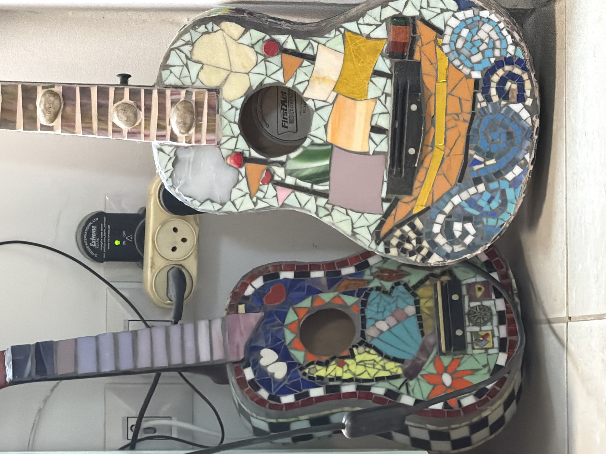The scene depicts two acoustic guitars, extensively decorated with mosaic tiles, leaning against a light-colored wall in an indoor setting in Tel-Aviv, Israel. The location is indicated by the presence of standard Israeli Type H electrical outlets and a "G-TRADE INTERNATIONAL LTD Extreme" surge protector visible in the background.
The left guitar features a vibrant, multi-patterned mosaic on its body. Notable elements include two white hearts and one red heart on a blue background near the top, an orange and green sunburst pattern surrounding the sound hole, a large light blue butterfly or fan-like shape, and an orange flower towards the bottom. The perimeter of its body is bordered by a black and white checkerboard mosaic. Its fretboard is adorned with mosaic tiles in shades of lavender and purple. A black strap or cable hangs down the left side of this guitar.
The right guitar, slightly larger, displays a thematic mosaic. The lower portion of its body depicts an orange sailing ship with yellow sails, situated on blue and white mosaic waves. The upper body features light green and yellow leaf-like patterns and red and orange flags near the sound hole, inside which the brand "FirstAct" is visible. The fretboard is covered in beige and brown mosaic tiles and has three oval-shaped, light-colored stones embedded, one of which bears the inscription "trust."
In the background, a white wall is visible with multiple electrical components. A white Type H electrical outlet is partially obscured on the left. To the right, a black "G-TRADE INTERNATIONAL LTD Extreme" surge protector, featuring a green "ON" indicator light, is plugged into another white Type H wall outlet. A light brown two-pin adapter is plugged into the surge protector, with a black electrical cable extending from it. Additional black cables are present behind and around the guitars and power outlets. The floor in the foreground is composed of light-colored tiles with a visible grout line running vertically. No people are present, and all objects are static.