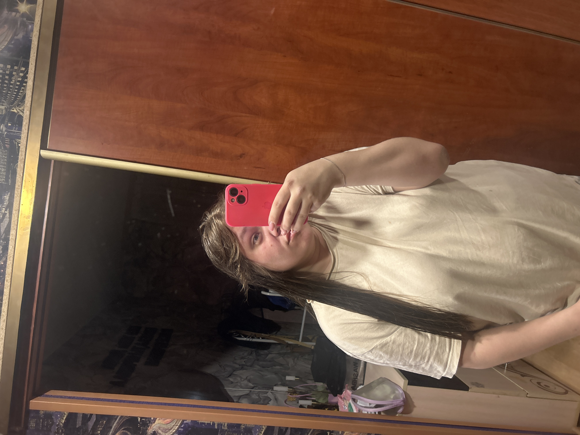A person with long dark hair is taking a selfie with a red phone in front of a mirror. The background shows a dark interior, possibly a room, with a wardrobe or cabinet visible behind the person. The mirror frame has a decorative pattern on the left and bottom. The location is Ольховка, Russia, but specific street or building details are unavailable from the image.