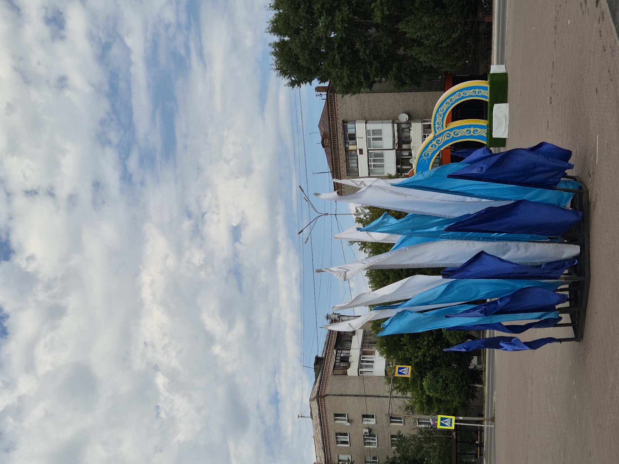 The image captures an outdoor scene with flags, buildings, trees, and a decorative archway. In the foreground, a cluster of blue and white flags are displayed on stands. Behind the flags, a multi-story brick building is visible, with windows and balconies. Adjacent to the building are several large green trees. To the right of the building, a decorative archway with yellow and blue intricate patterns is present, situated on a patch of artificial grass. Two pedestrian crossing signs are also visible. The sky is partly cloudy. The location is identified as Kokshetau, Kazakhstan.