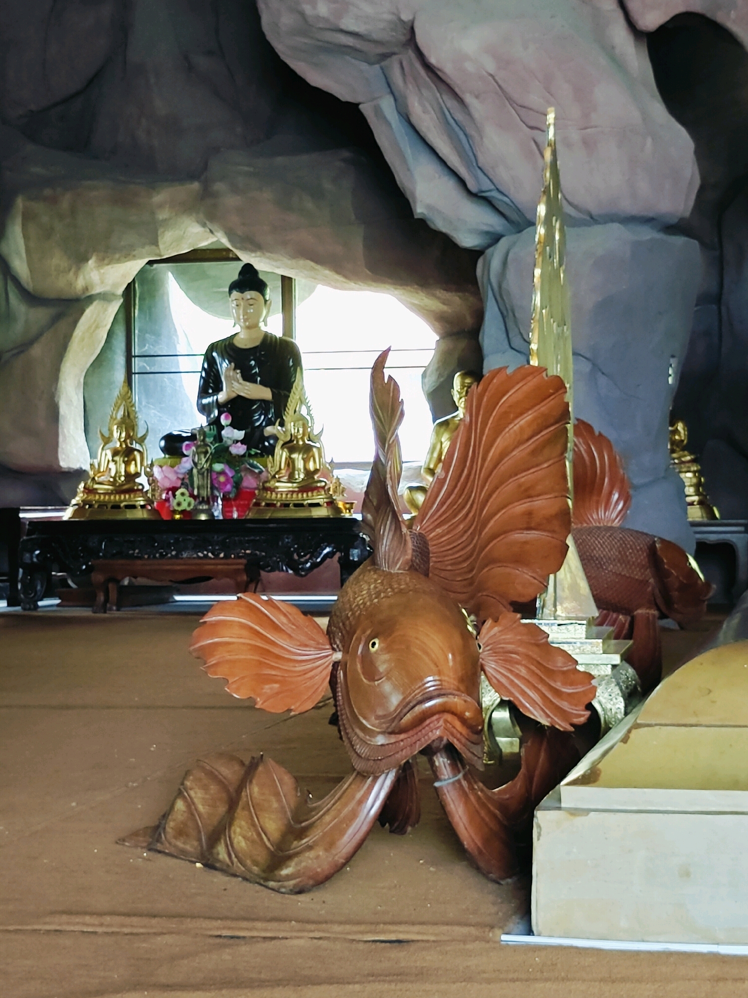 This is a photograph taken inside a cave-like structure with rough, textured rock formations overhead. In the foreground, a large, intricately carved wooden fish sculpture is positioned on the floor, its body and fins detailed and spread out. Behind the fish, a dark wooden table or altar is visible, adorned with golden Buddha statues and a floral arrangement. A larger Buddha statue in dark robes, with hands clasped in a prayer gesture, is seated behind the altar, illuminated by natural light filtering in from a window or opening behind it. The floor appears to be covered in a brown, sandy material. The scene suggests a Buddhist shrine or temple, possibly located in the Hantra Subdistrict of Thailand, given the cultural elements depicted.