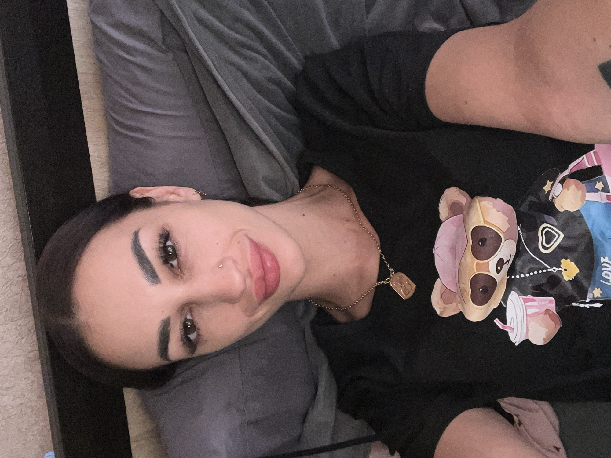 The image shows a woman lying on a bed, looking at the camera. She has dark hair pulled back, dark eyebrows, and is wearing makeup. She has a nose ring and hoop earrings. She is wearing a black t-shirt with a graphic of a cartoon bear holding a drink. She is wearing a gold necklace with a rectangular pendant. The background appears to be a bedroom, with a textured wall and a gray blanket. The image was taken in Bashtanka, Ukraine.