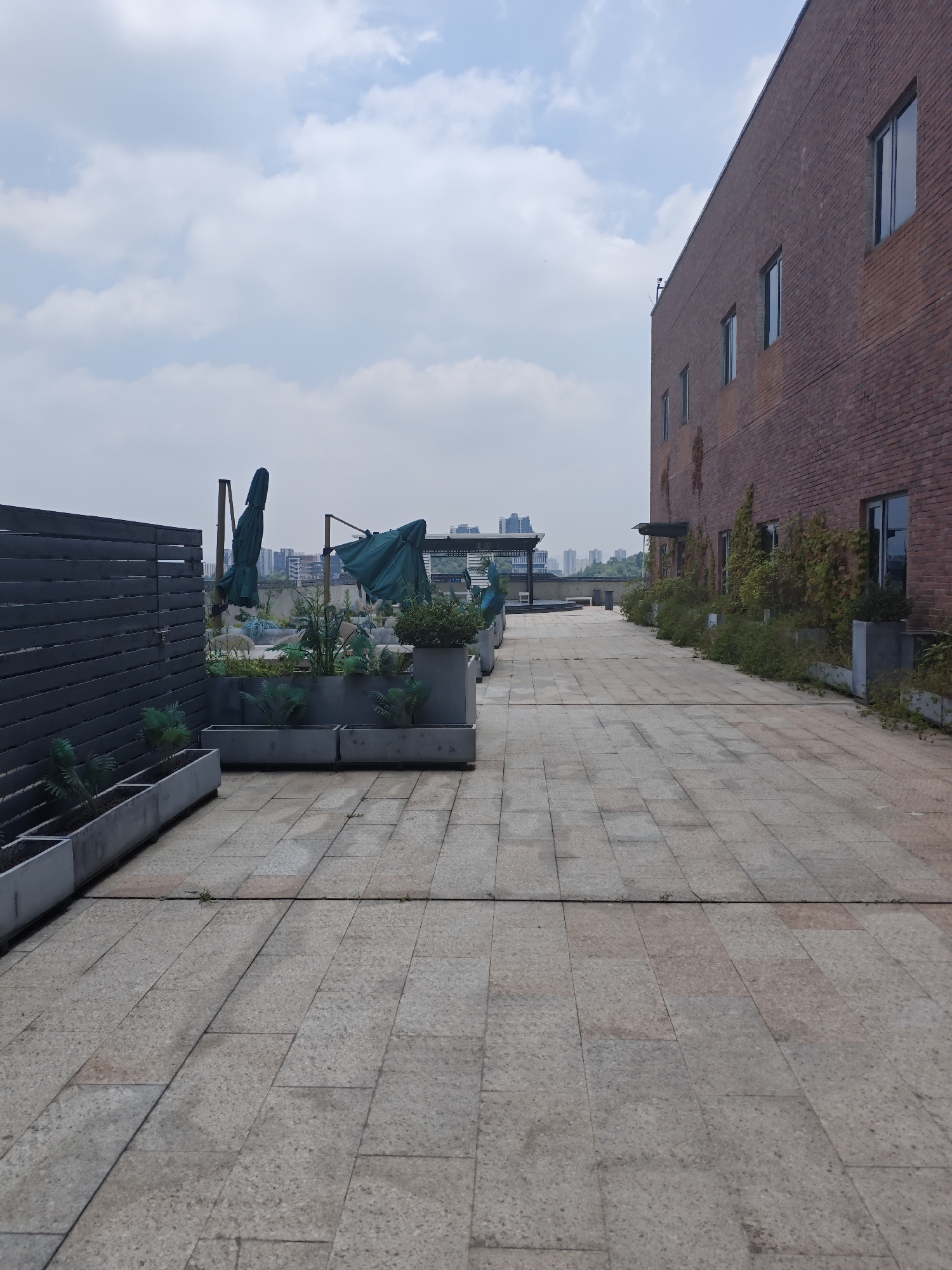 An exterior shot captures a rooftop terrace in Chongqing High-tech Zone, China. The terrace features a brick building on the right, with several windows and vines growing on its facade. A paved walkway extends from the foreground into the distance, lined with planters containing greenery and several covered green umbrellas. A dark wooden fence is visible on the left side of the frame. The sky is partly cloudy.